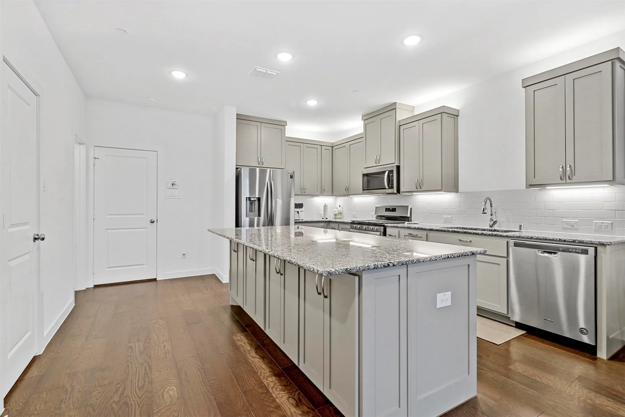 Spacious kitchen featuring an extended island with granite countertops, stainless steel appliances, light gray shaker cabinetry, a white subway tile backsplash, and dark wood-finish flooring