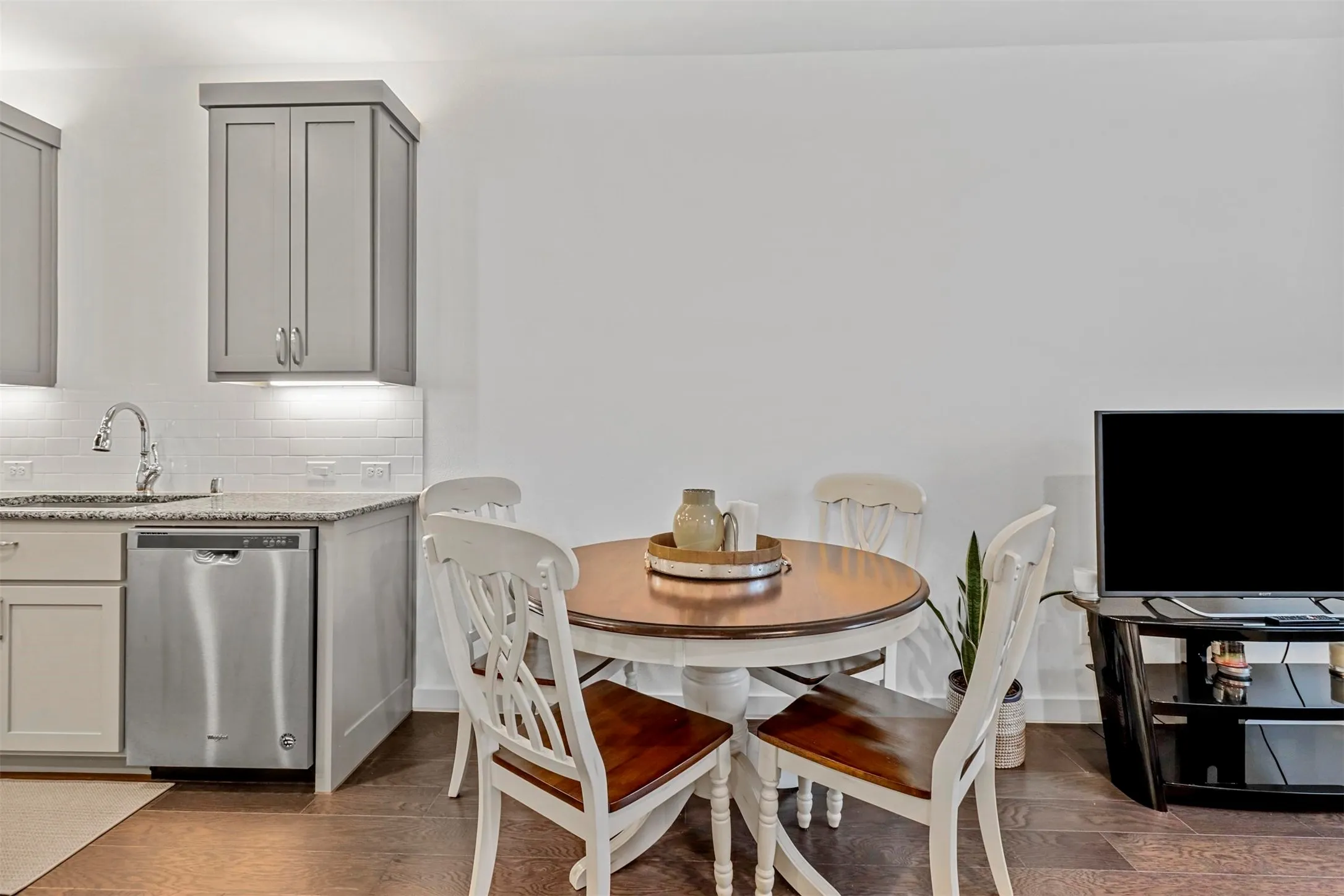 Kitchen featuring wood flooring, gray shaker-style cabinetry, stainless steel dishwasher, white subway tile backsplash, and light-toned countertops.