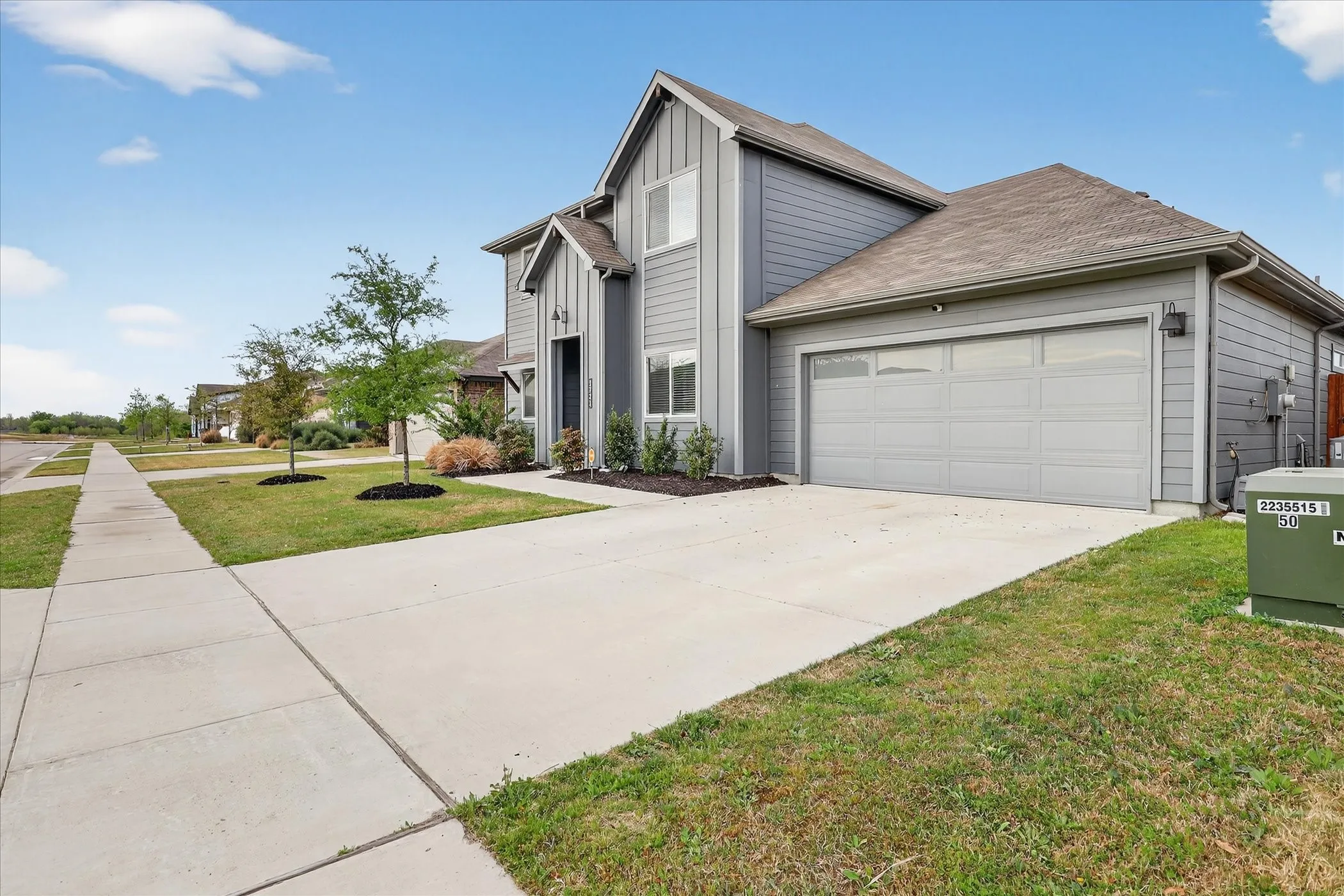 Contemporary gray siding exterior with a contrasting dark front door, attached garage with frosted glass panels, concrete driveway, and a manicured lawn with young trees