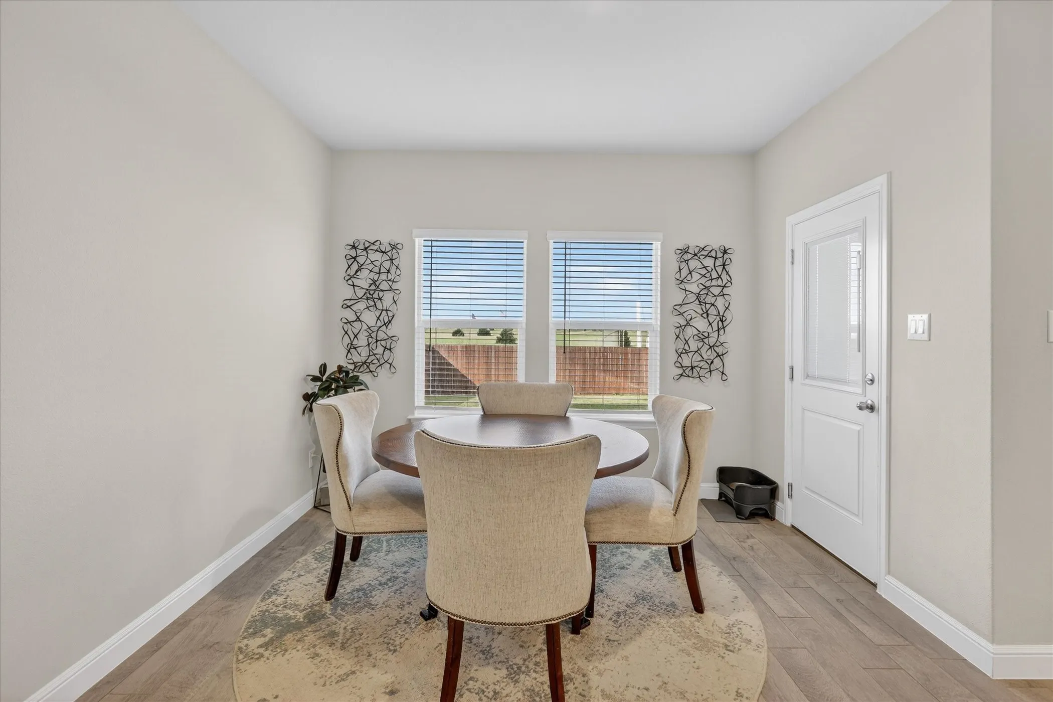 Bright dining area featuring wood-finish flooring, two large windows with horizontal blinds, and a white paneled door with a frosted glass insert