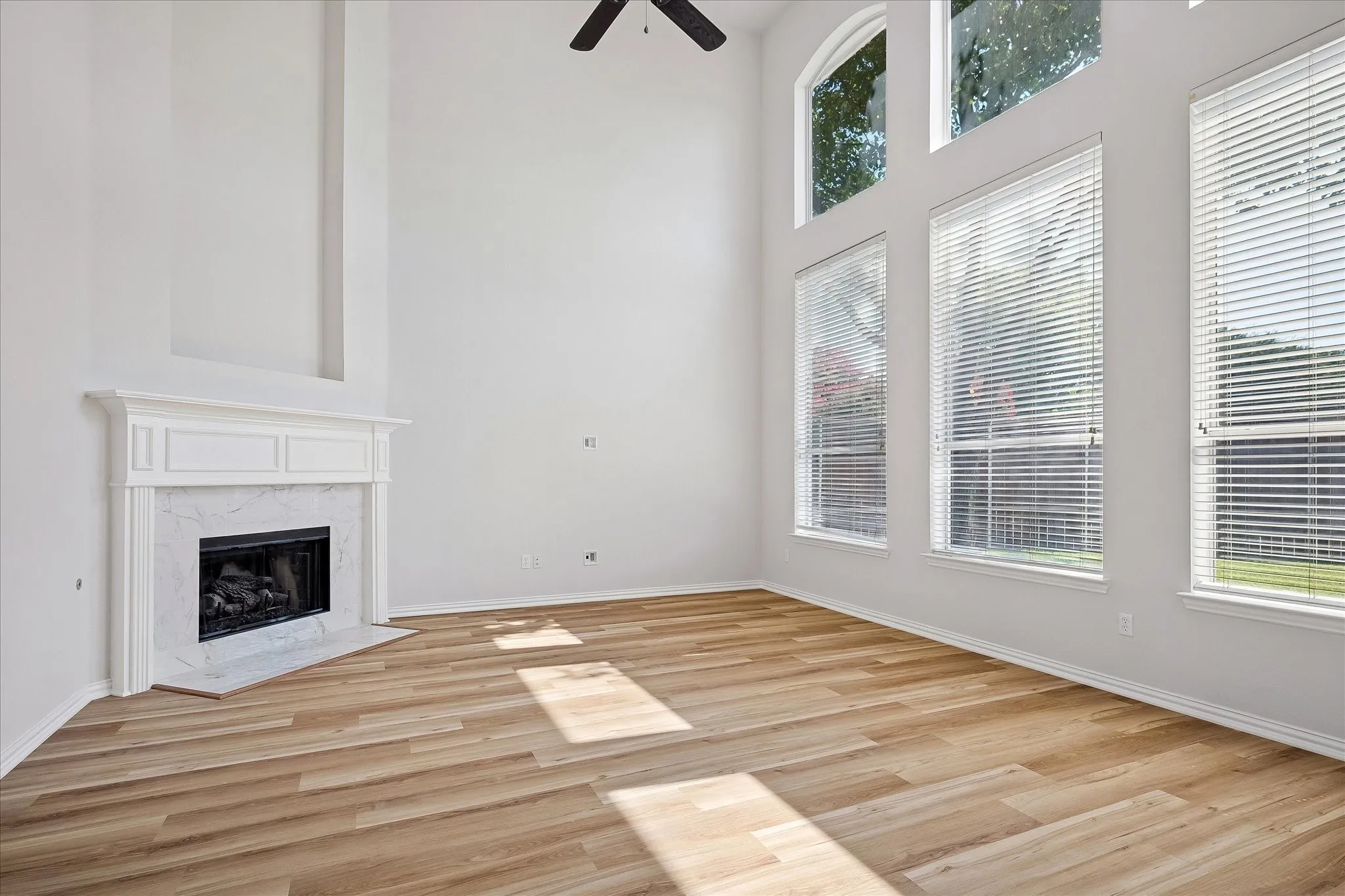 Two-story living area featuring a fireplace with a white mantel and marble surround, wood-finish flooring, and multiple tall windows with blinds
