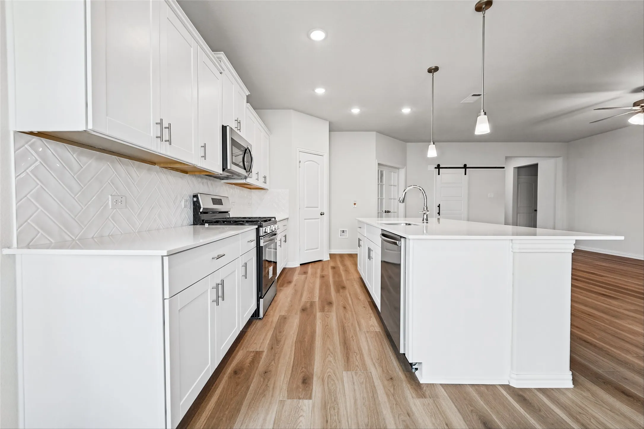 Kitchen featuring white shaker cabinetry, stainless steel appliances, a white tile herringbone backsplash, a large center island with a sink, and wood-finish flooring