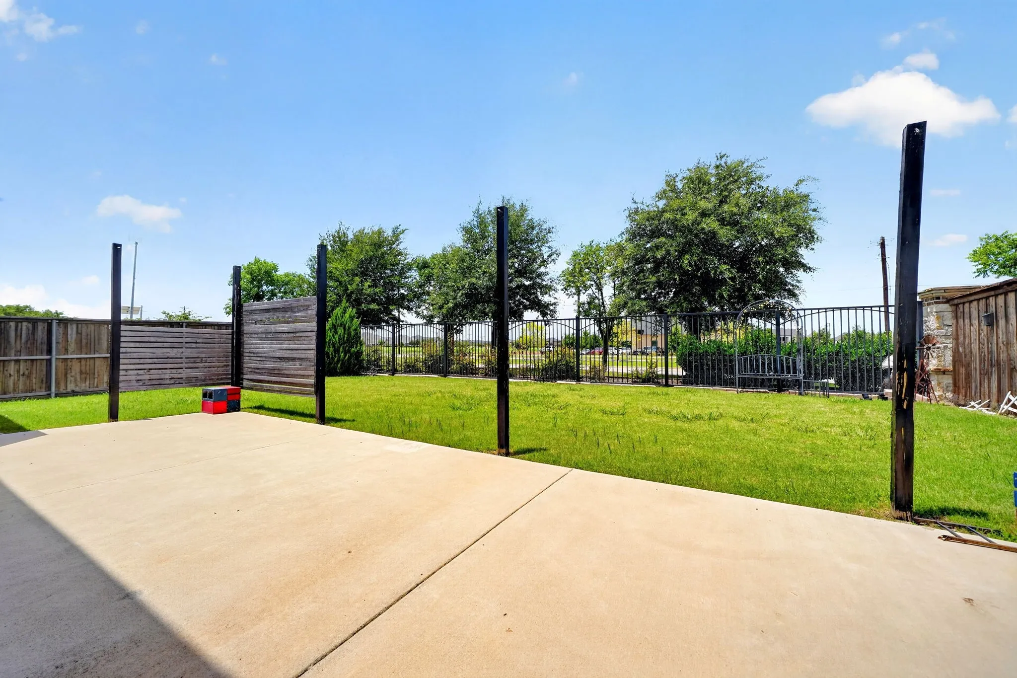 Expansive concrete patio with a spacious grassy area, wood privacy fencing, and black metal fencing with mature trees