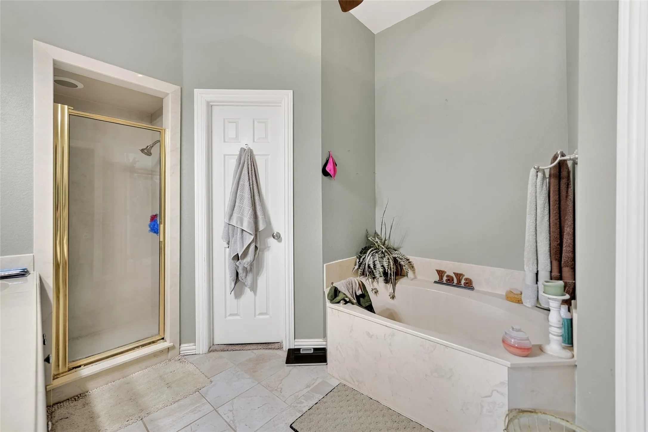 Bathroom featuring a built-in tub with a light-toned surround, an enclosed shower with a gold-toned frame, and light-colored tile flooring