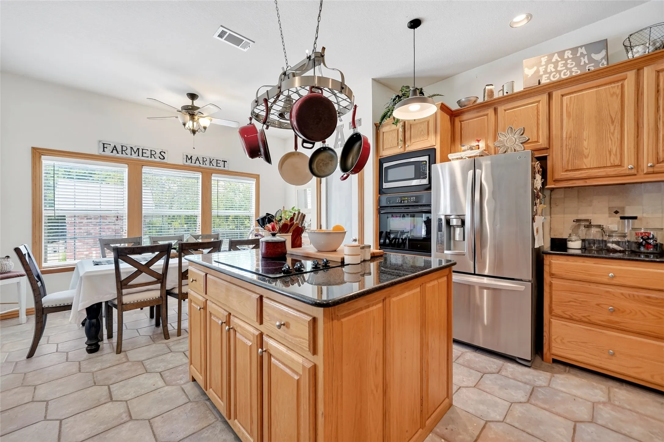Spacious kitchen featuring an island with a black countertop, stainless steel appliances, light wood cabinetry, and a pot rack