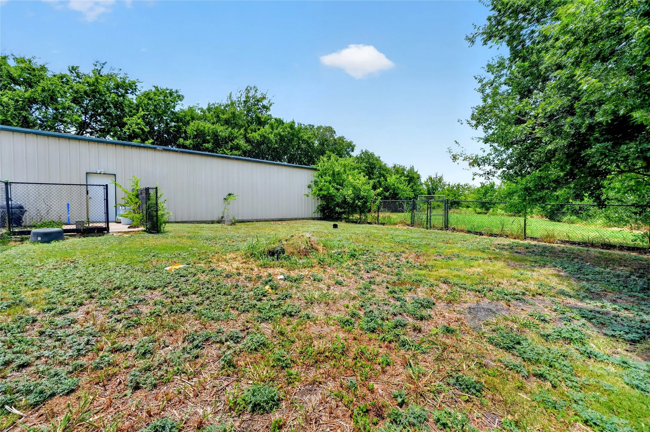 Expansive outdoor area featuring a metal-sided building with a pedestrian entry door, chain-link fencing, and mature trees bordering the property
