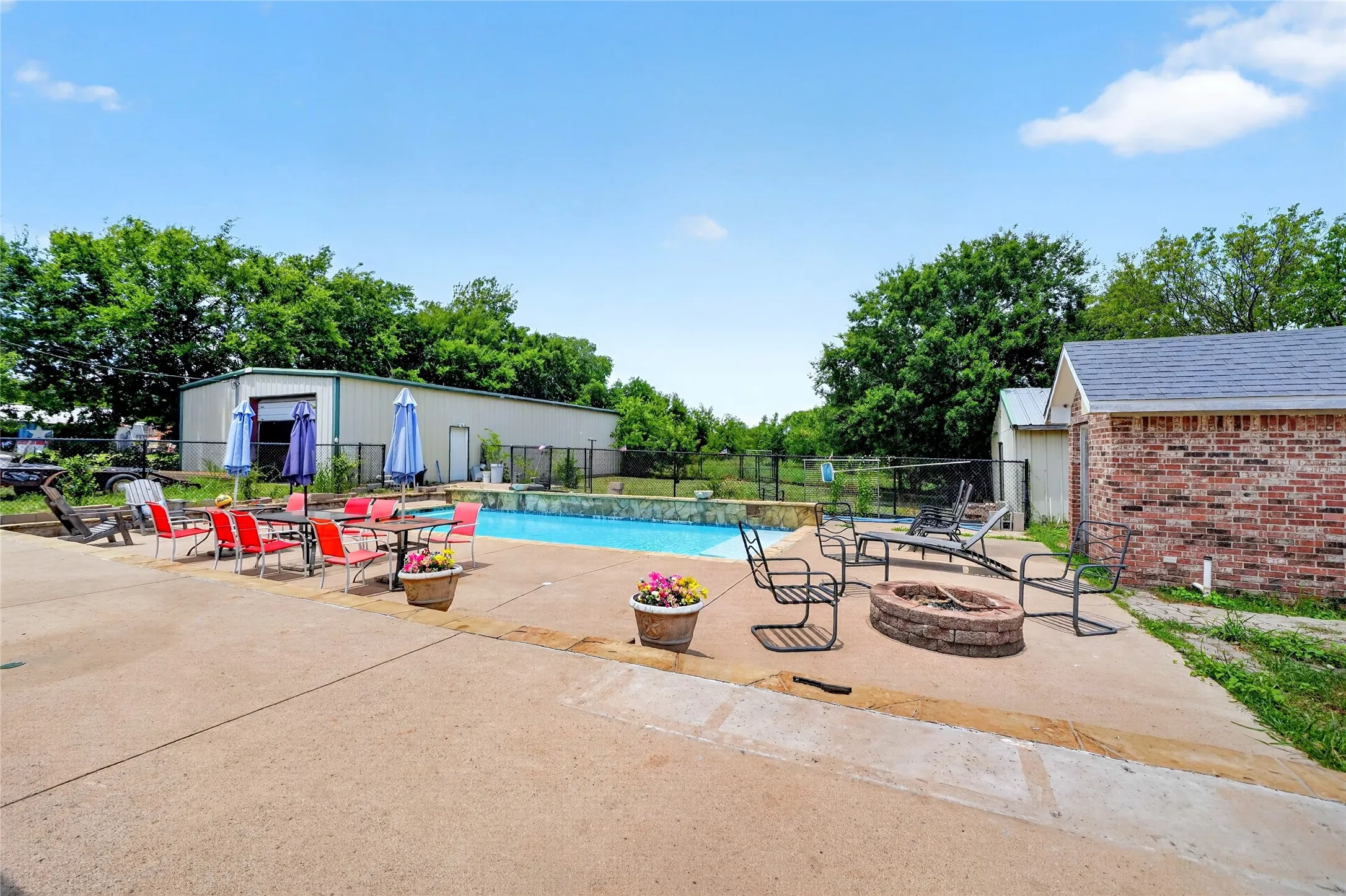 Outdoor concrete patio featuring a built-in fire pit, a fenced swimming pool, and a brick-sided outbuilding with a shingled roof