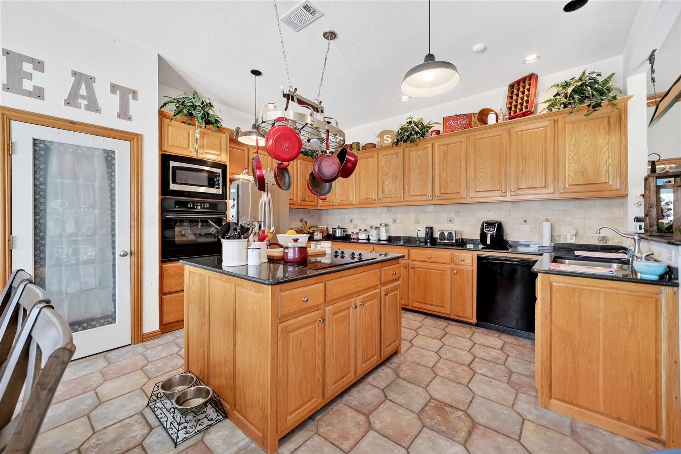 Kitchen featuring honey-toned cabinetry, a central island with a cooktop, stone tile flooring, black countertops, and built-in double ovens