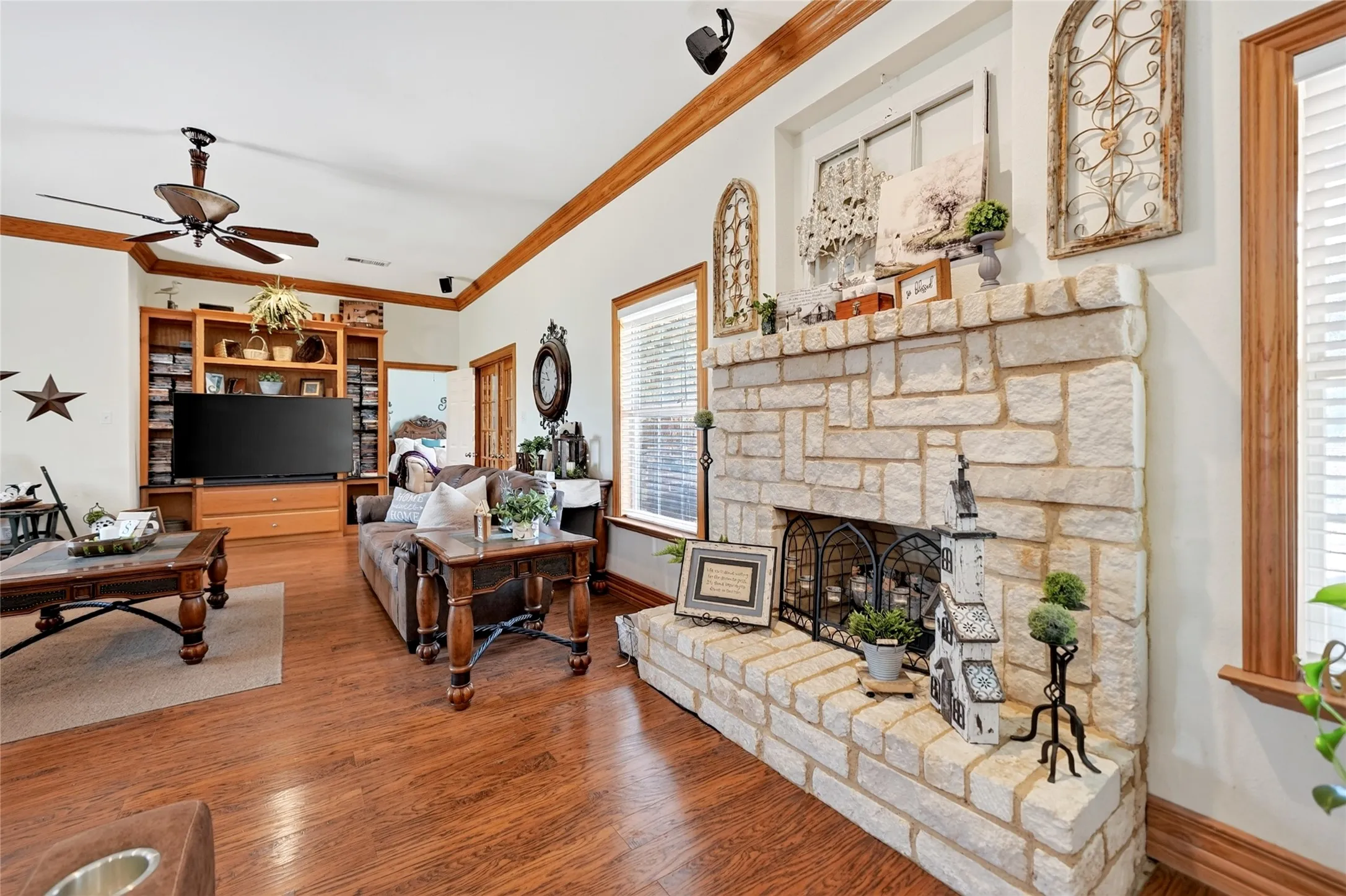 Stone-clad fireplace with hearth, wood-finish flooring, built-in media center, ceiling fan, and crown molding