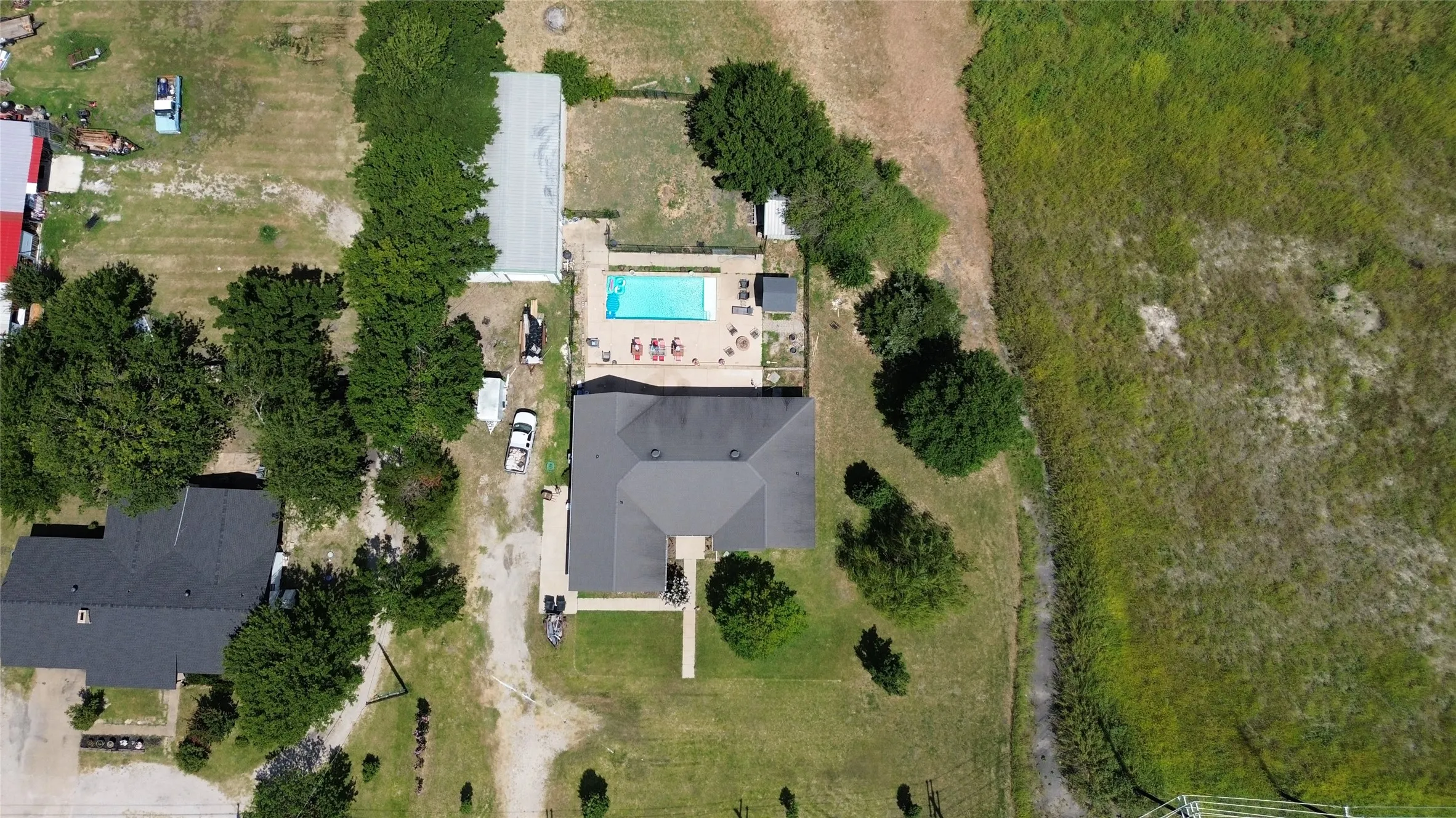 Single-story property with a dark shingle roof, featuring a rectangular swimming pool with a surrounding patio, a lengthy gravel driveway, and a separate outbuilding with a metal roof