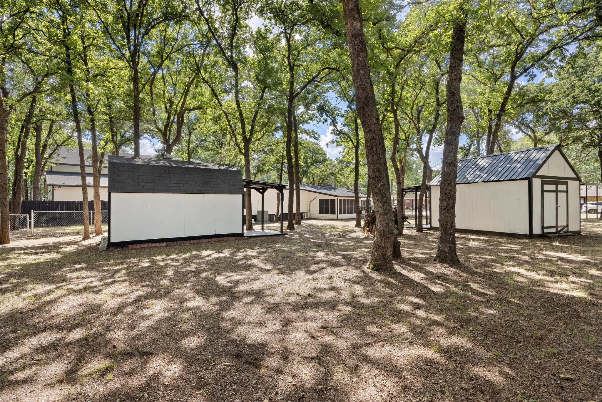 4617 Lyndi Ln
Granbury, TX  76049 Expansive treed lot featuring two detached storage sheds with white siding and dark trim, and a separate structure with an attached covered porch