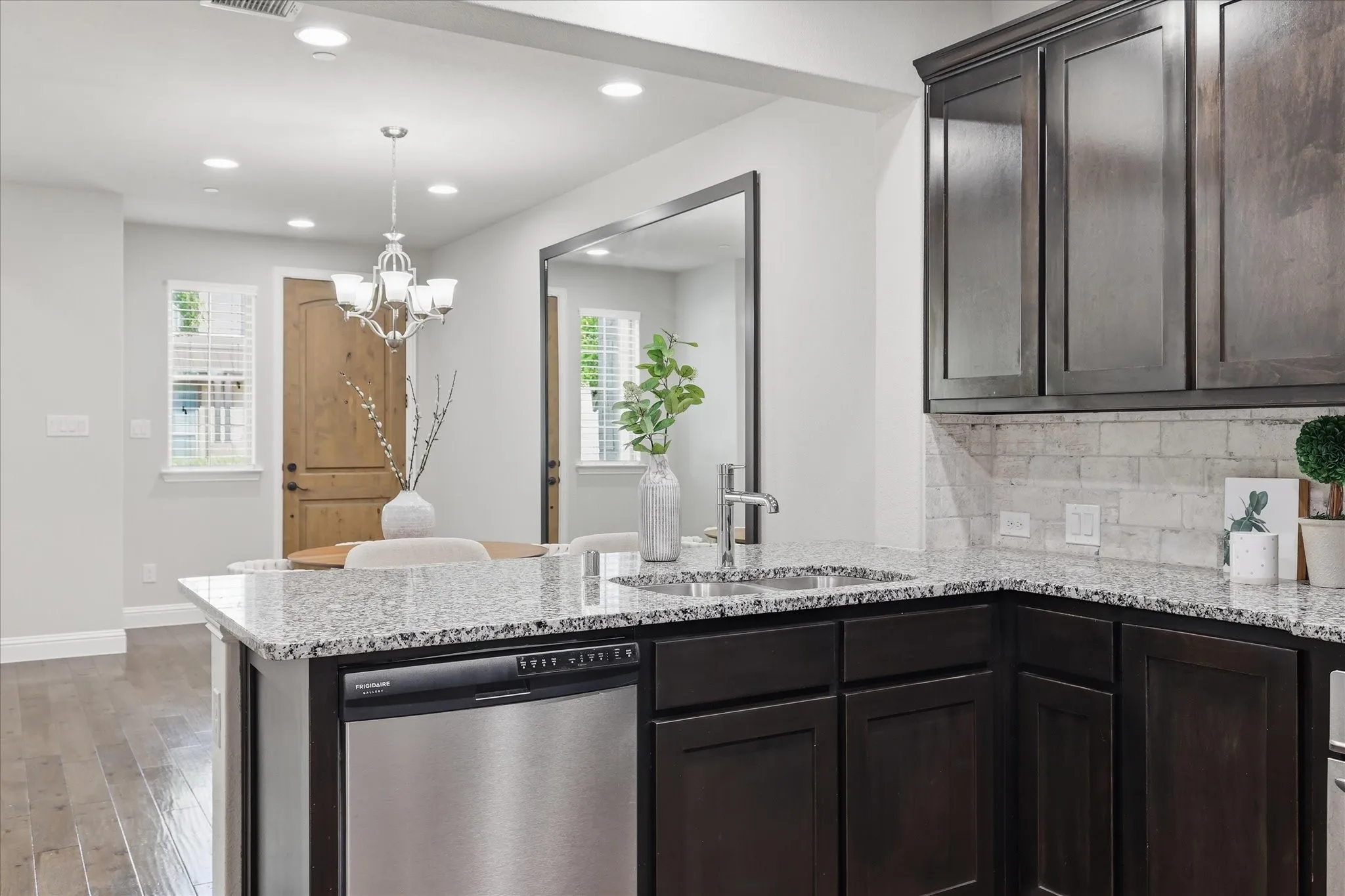 Kitchen featuring dark wood-finish cabinetry, light-toned granite countertops, a stainless steel dishwasher, and a tile backsplash