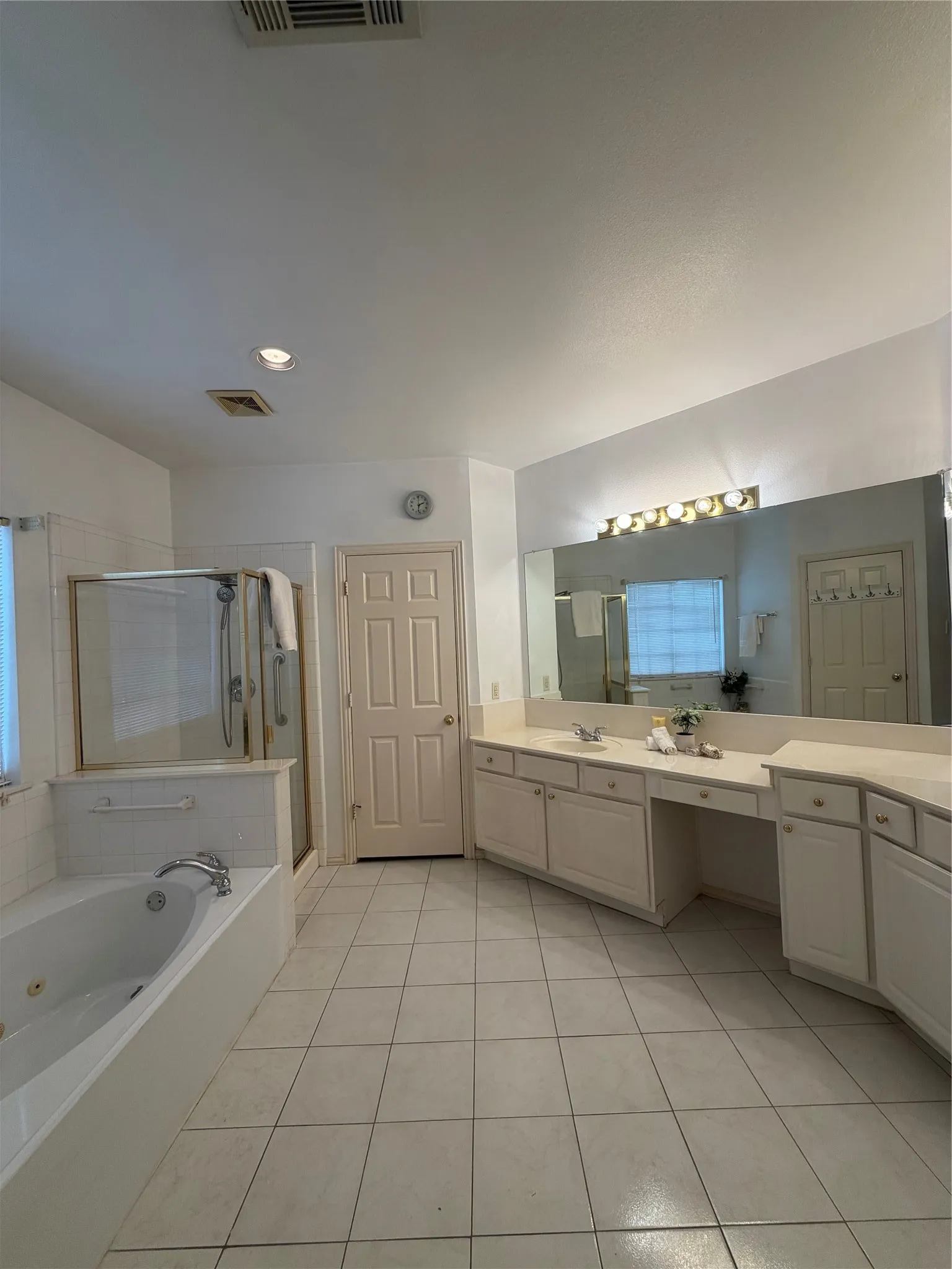 Spacious bathroom with white tile flooring, dual vanities, large mirror, jetted soaking tub, and glass-enclosed shower