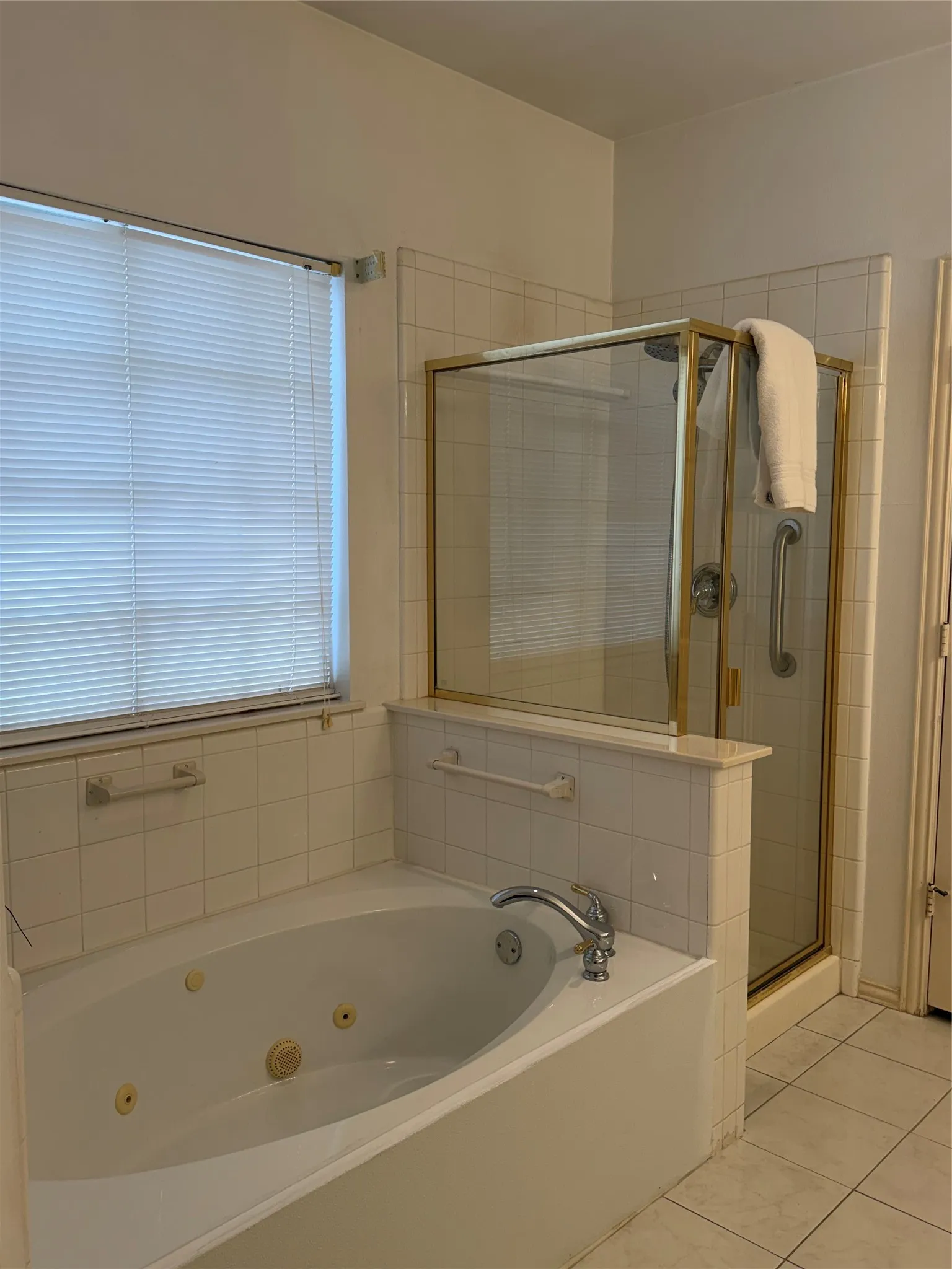 Soaking tub and enclosed shower with gold-tone framing, complemented by white tile surrounds and a window with horizontal blinds