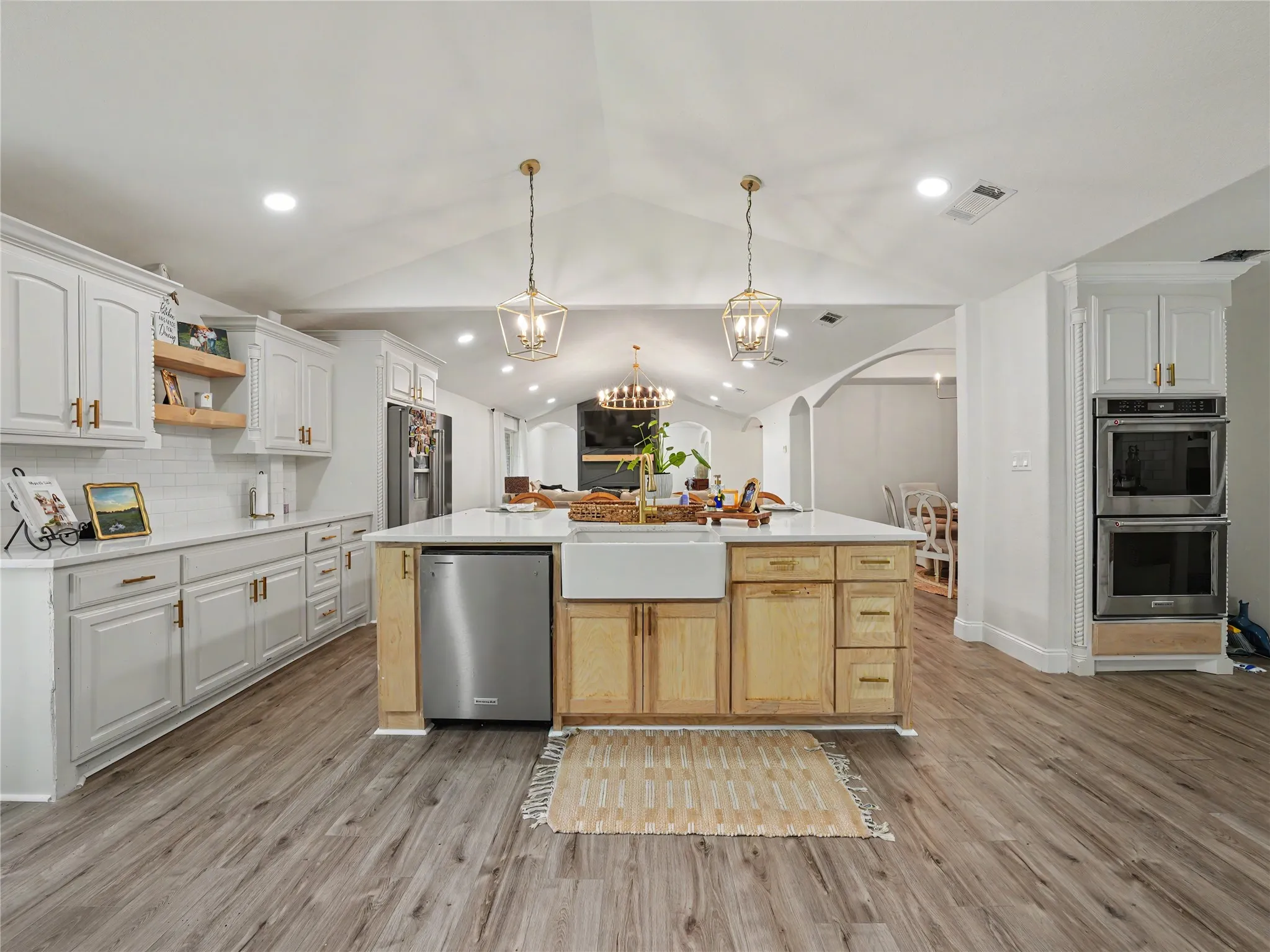 Spacious kitchen featuring an expansive center island, farm sink, light wood-finish flooring, white cabinetry with gold-tone hardware, and double wall ovens