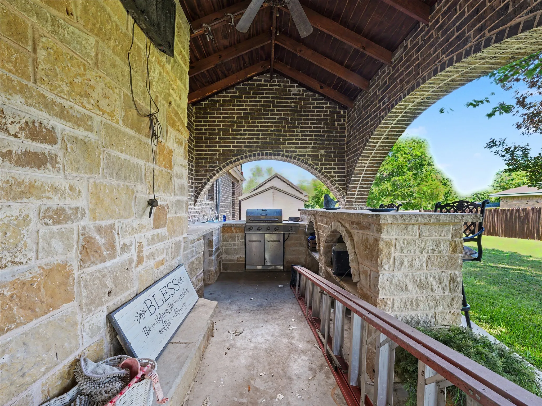 Covered outdoor kitchen featuring stone construction, brick archways, a wood-plank ceiling, and a built-in grill