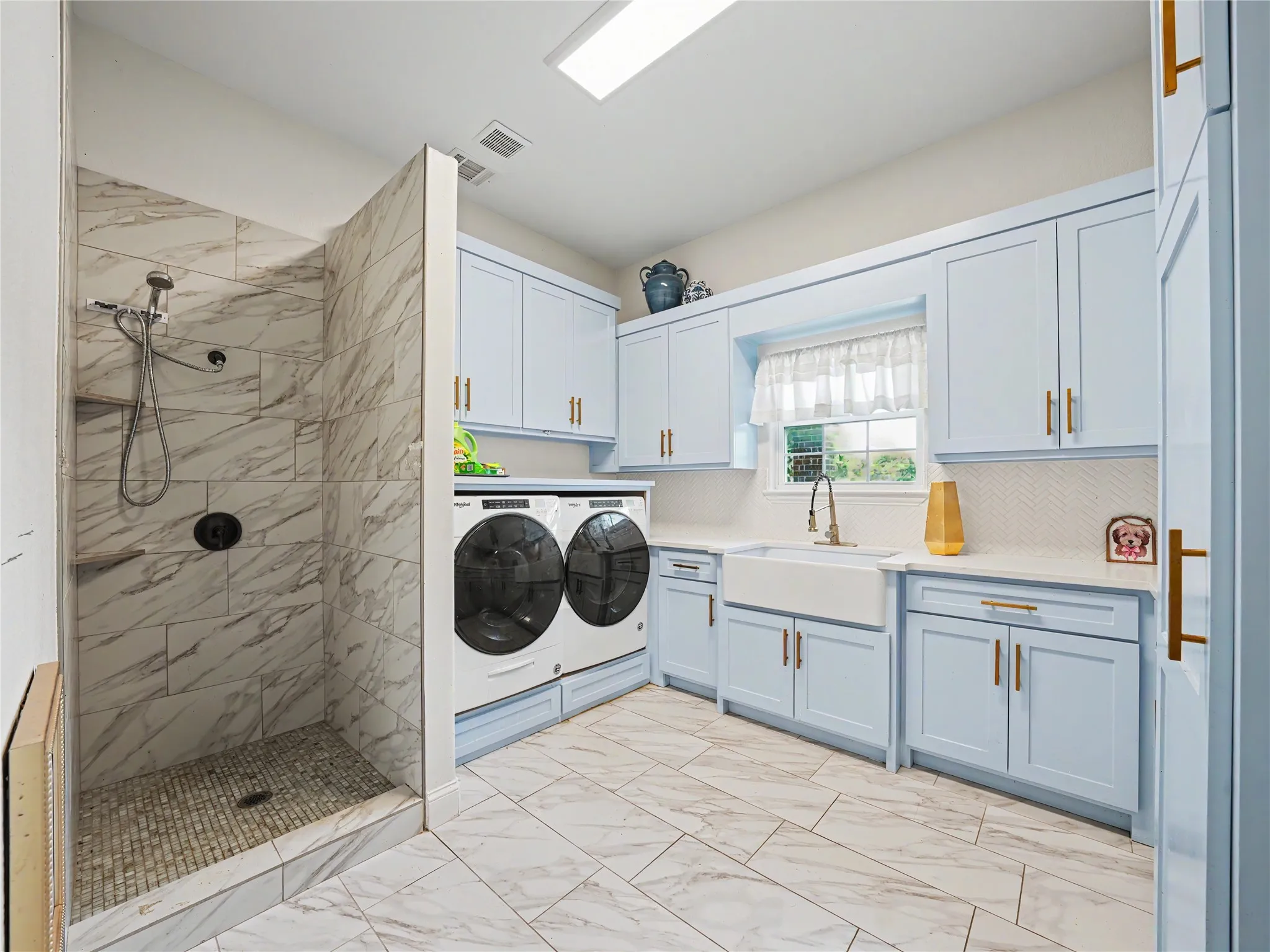 Utility room featuring a walk-in shower with stone tile, a white farmhouse sink with a gooseneck faucet, light blue cabinetry with gold-toned hardware, white countertops, and wood-finish tile flooring