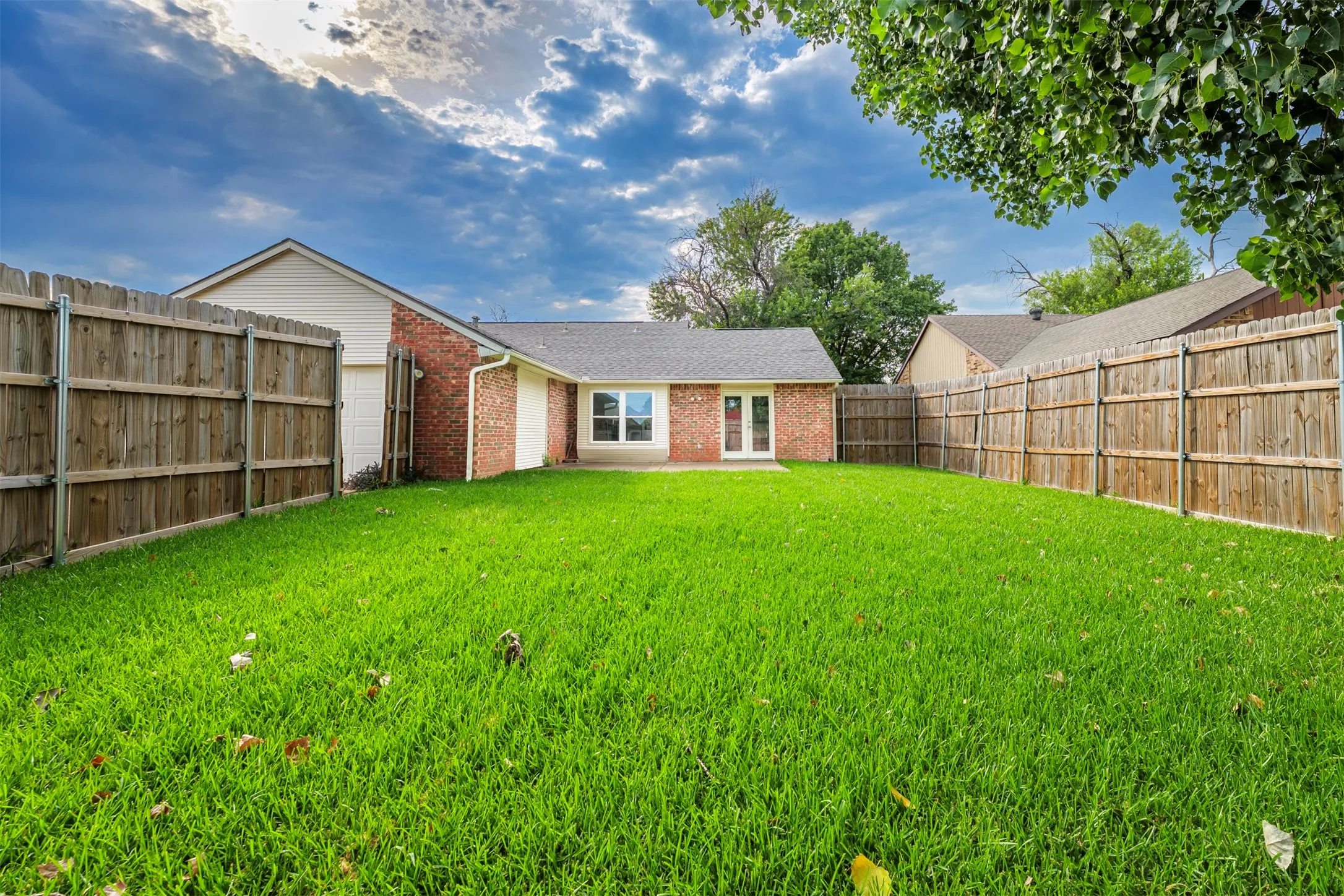 Expansive fenced backyard featuring a verdant lawn, brick exterior, double French doors, and a window with white trim