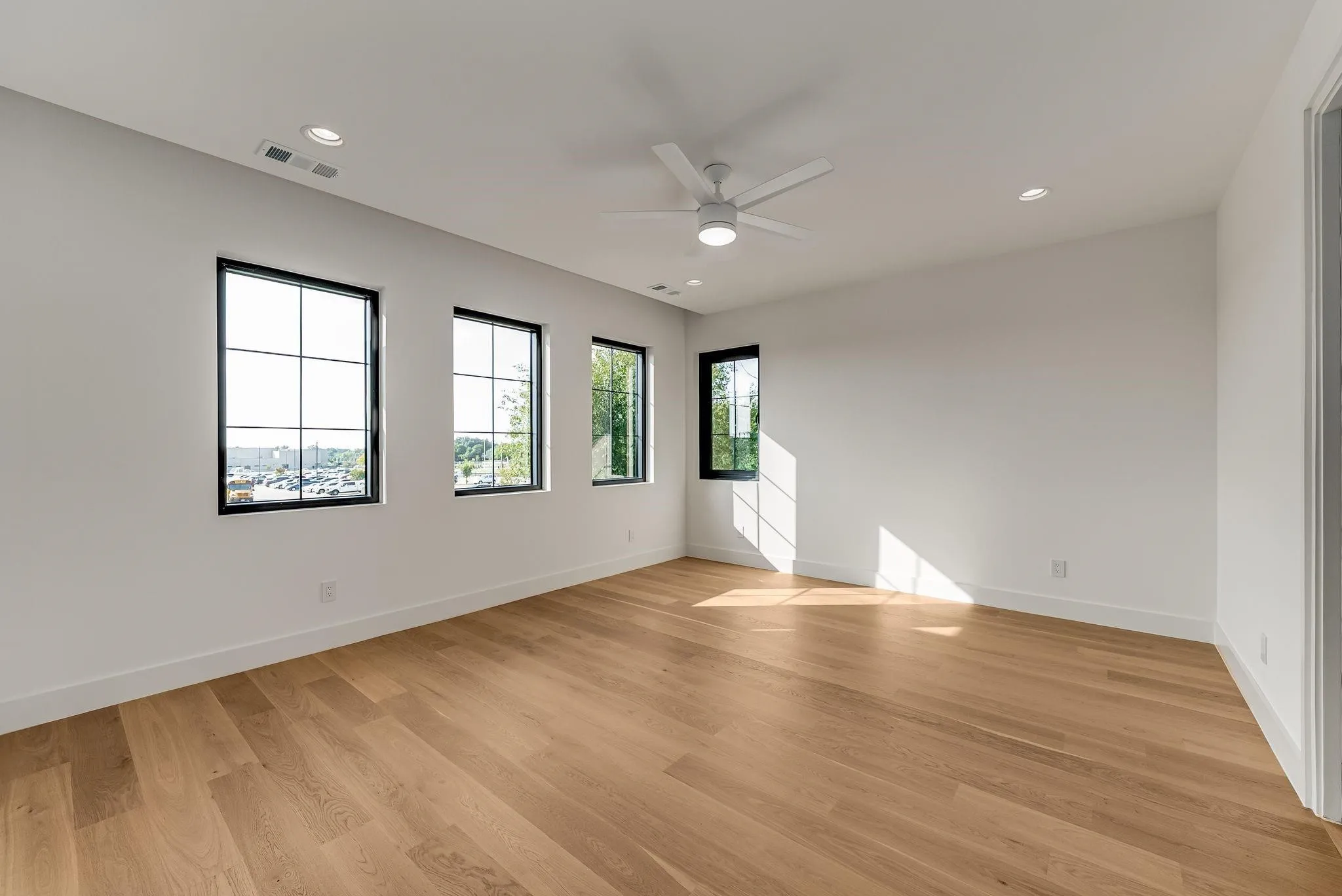 Spacious room featuring light wood-finish flooring, three large windows with black grilles, recessed lighting, and a ceiling fan