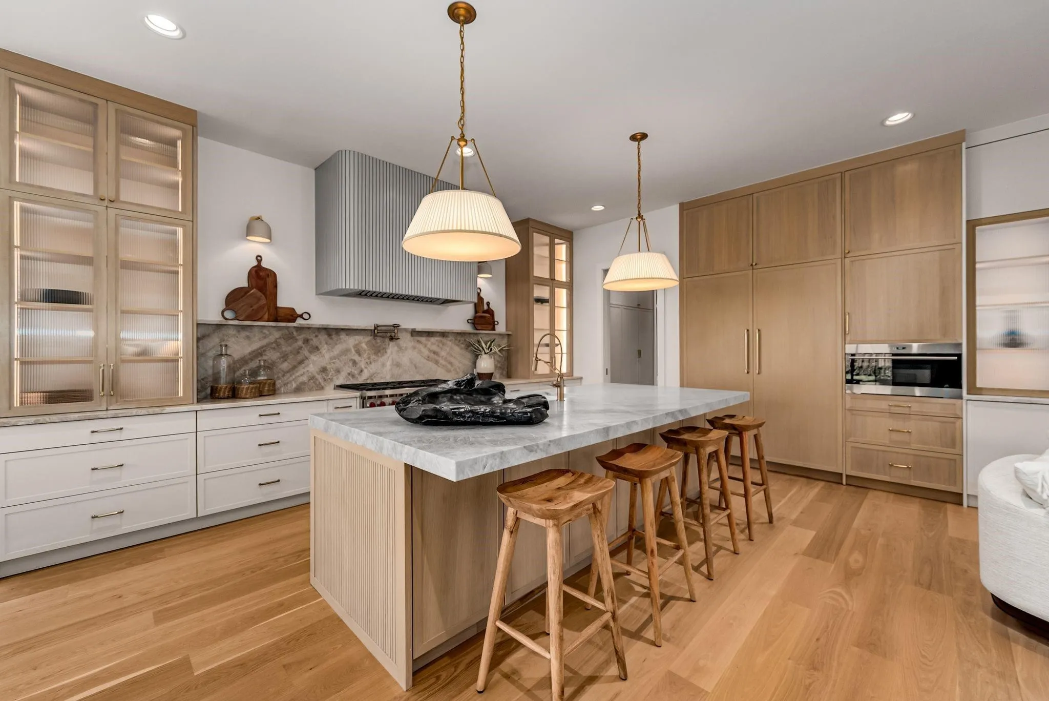 Open-concept kitchen featuring an oversized island with seating, natural wood-finish flooring, integrated appliances, a fluted range hood, and illuminated glass-front cabinetry