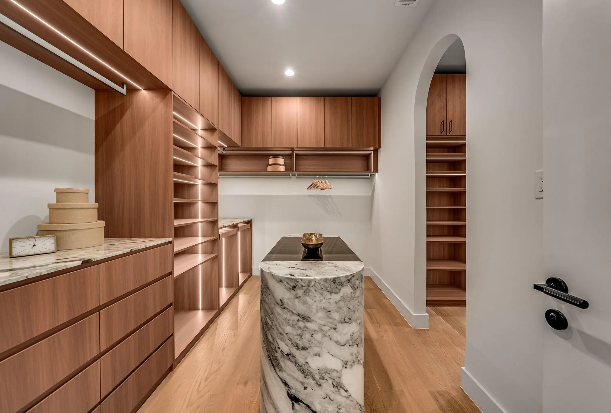 Custom closet system featuring wood-finish cabinetry, integrated lighting, marble-finish countertops, a central marble-clad island with dark countertop, and an arched entryway with additional shelving