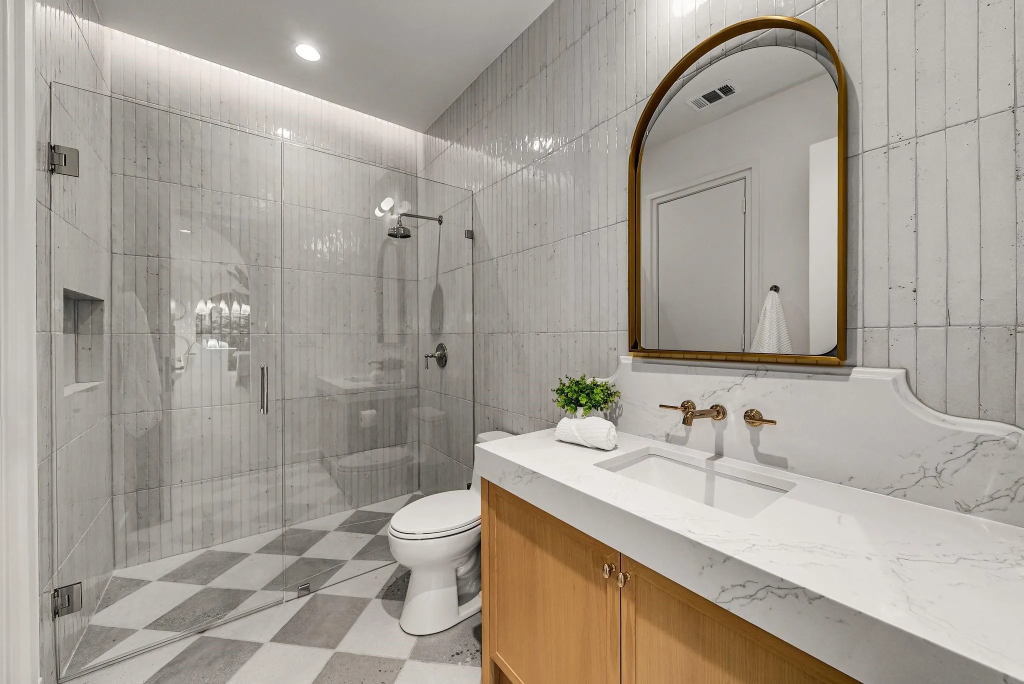 Bathroom featuring a glass-enclosed shower with tiled walls and a built-in niche, a checkered floor, a modern vanity with a light wood finish, a white countertop with integrated sink, and an arched mirror with a brass frame