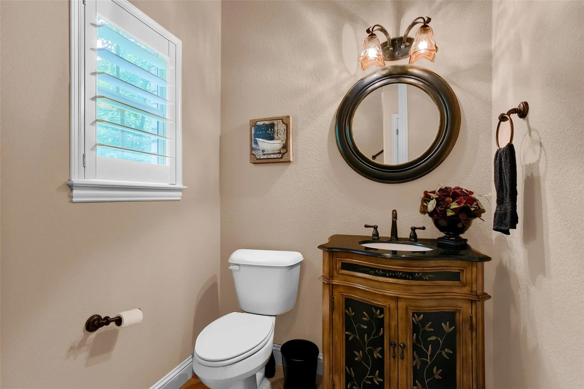 Powder room featuring a decorative vanity with a dark countertop and integrated sink, a round framed mirror, a two-light wall sconce, and a window with white plantation shutters