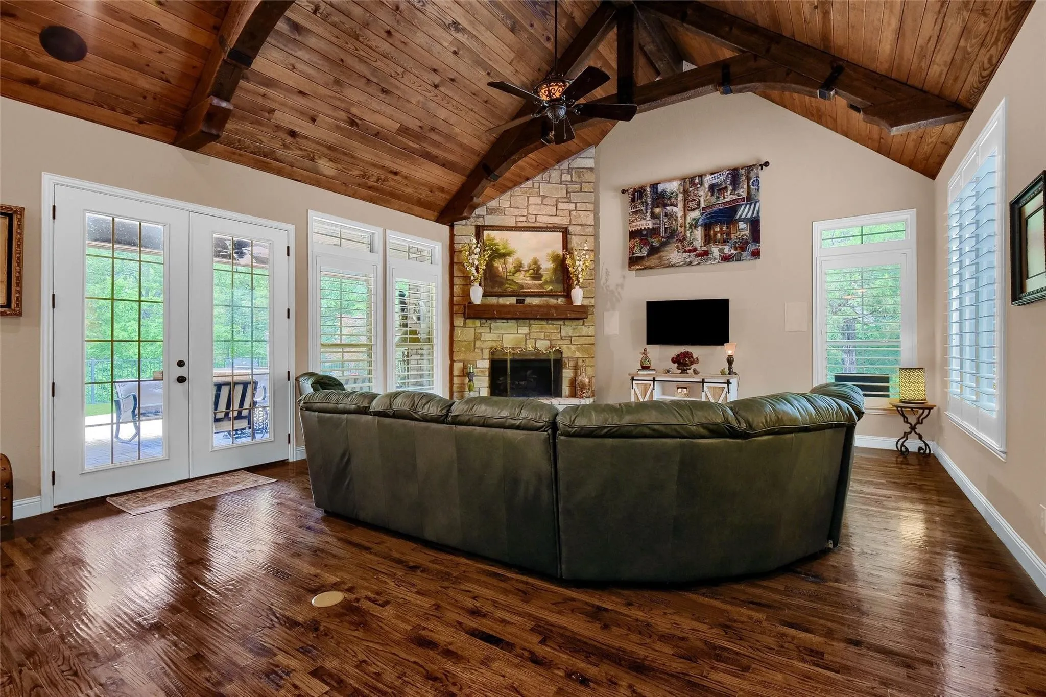 Spacious living area featuring a vaulted wood-paneled ceiling with exposed beams, rich wood-finish flooring, and a floor-to-ceiling stone fireplace