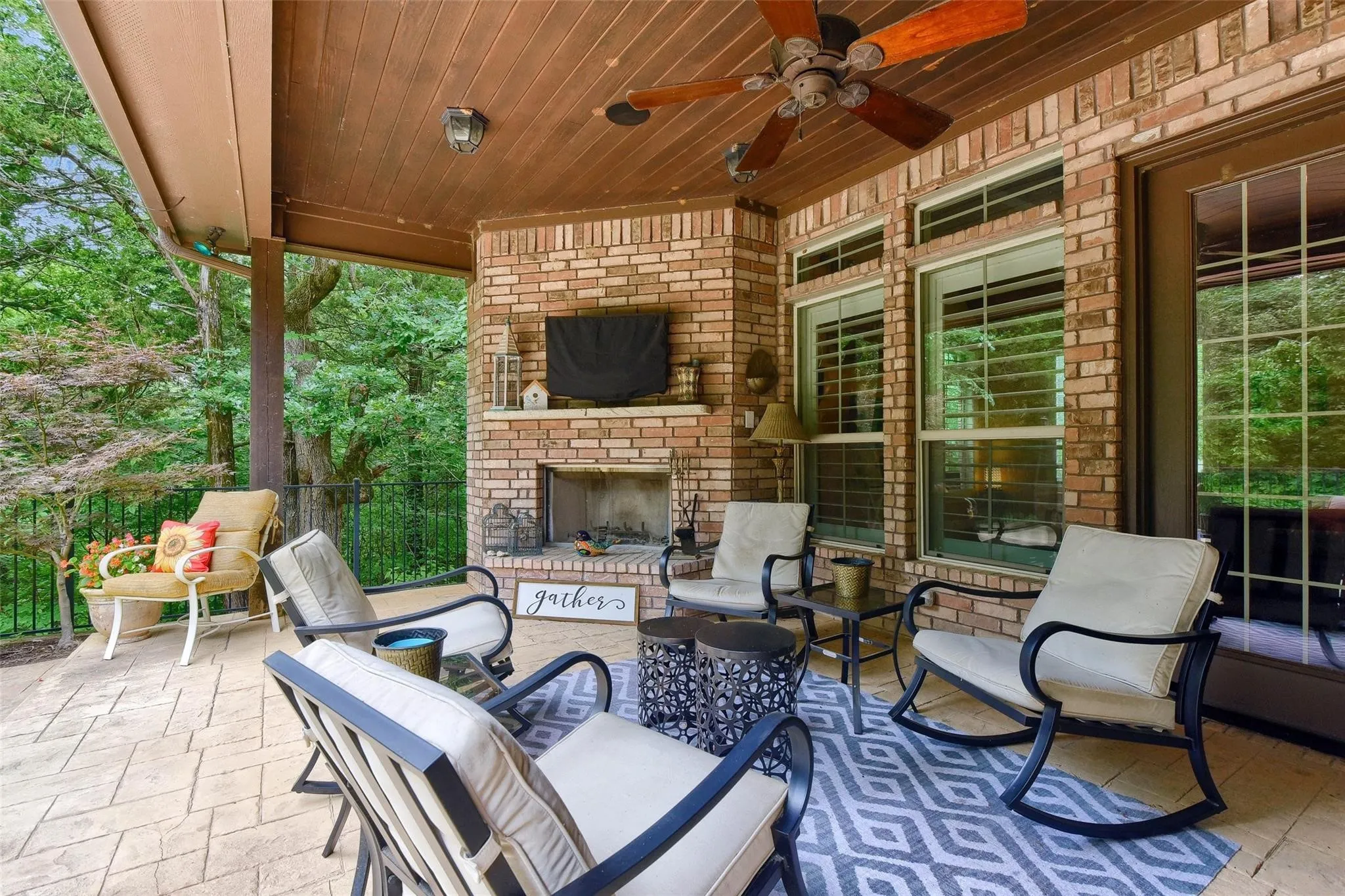 Expansive covered patio featuring a brick fireplace, wood-plank ceiling with a ceiling fan, patterned concrete flooring, and multiple windows with horizontal blinds