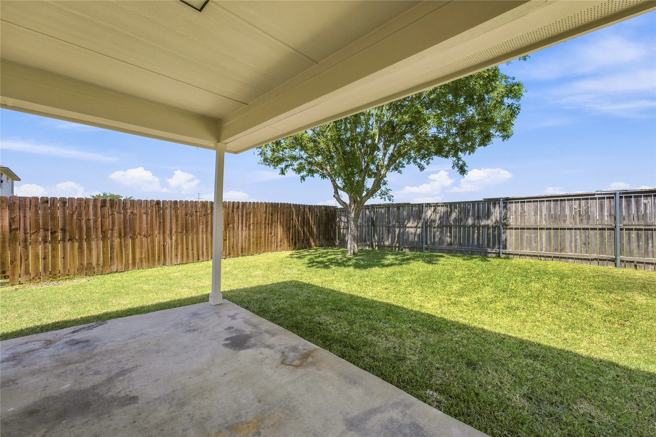 Covered concrete patio overlooking a private backyard with a mature tree, wood fencing, and manicured lawn
