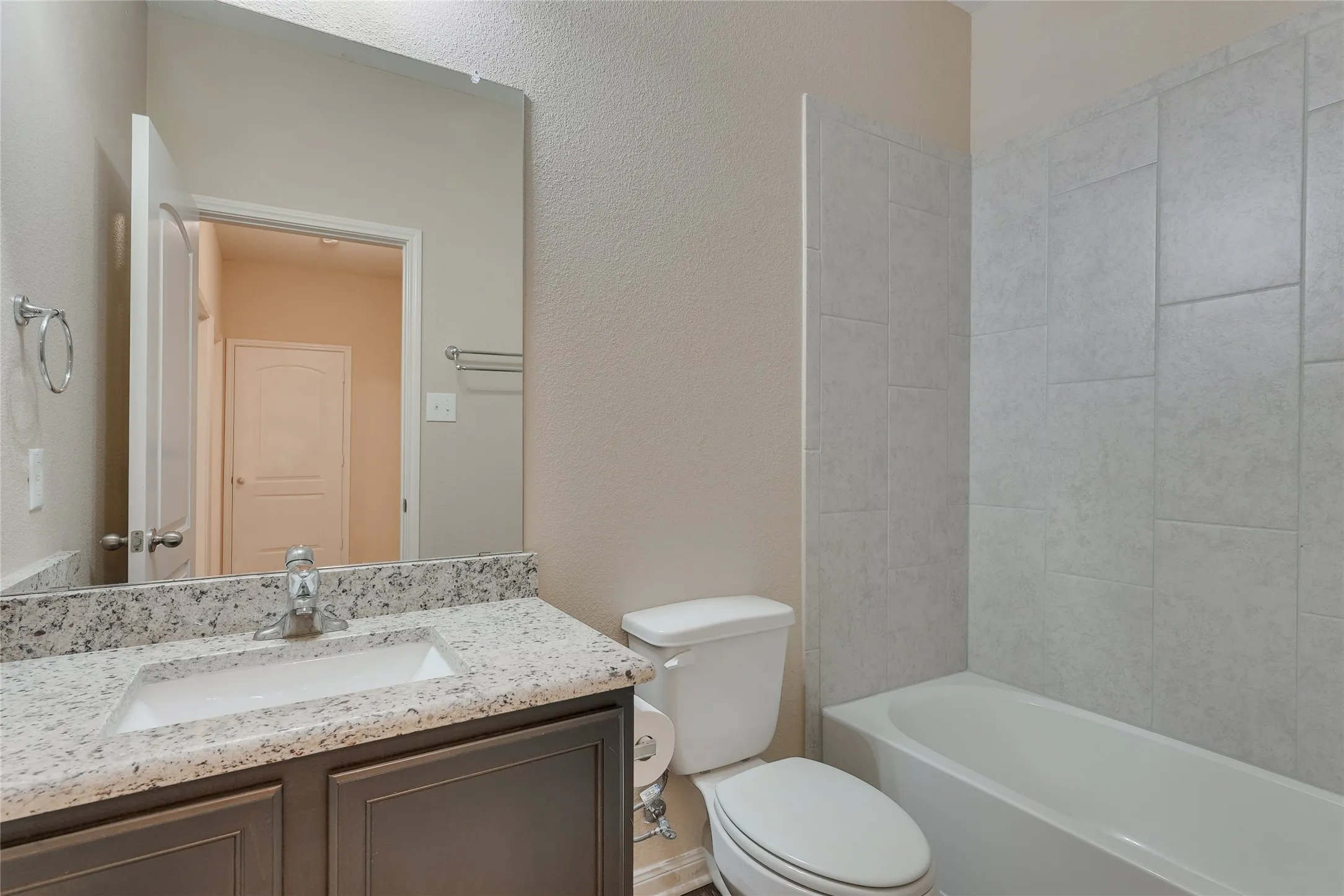 Bathroom featuring a vanity with a white rectangular sink, light-toned speckled countertop, dark wood-finish cabinetry, a white toilet, and a white bathtub with light gray tile surround