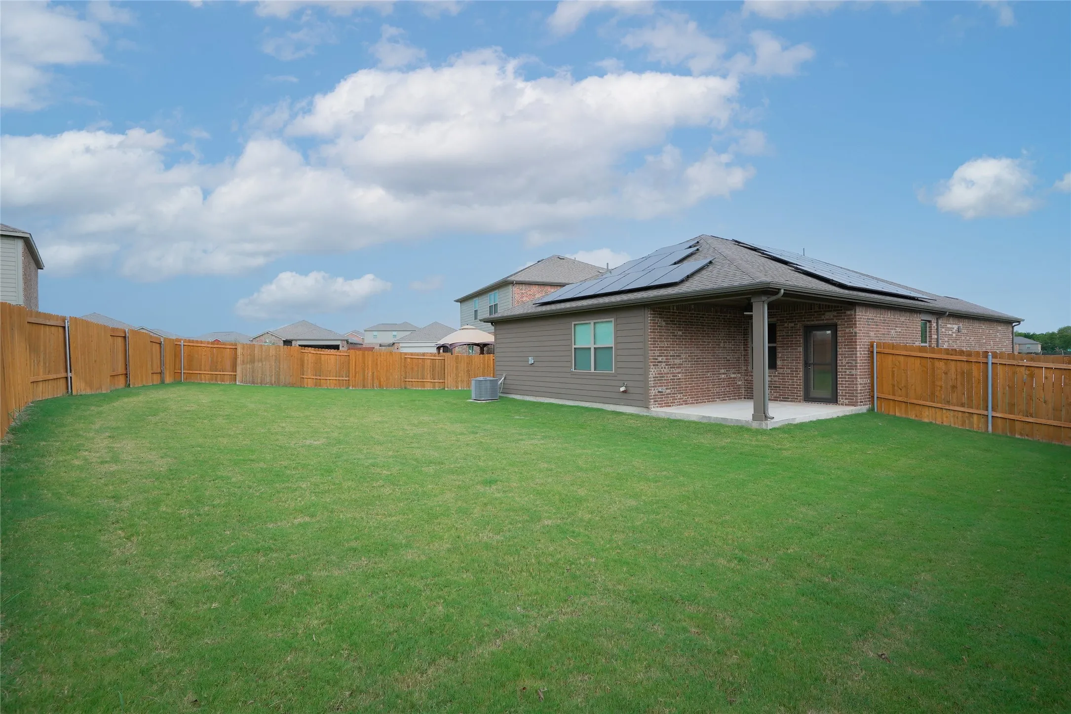 Expansive fenced backyard featuring a covered patio, brick and siding exterior, and roof-mounted solar panels