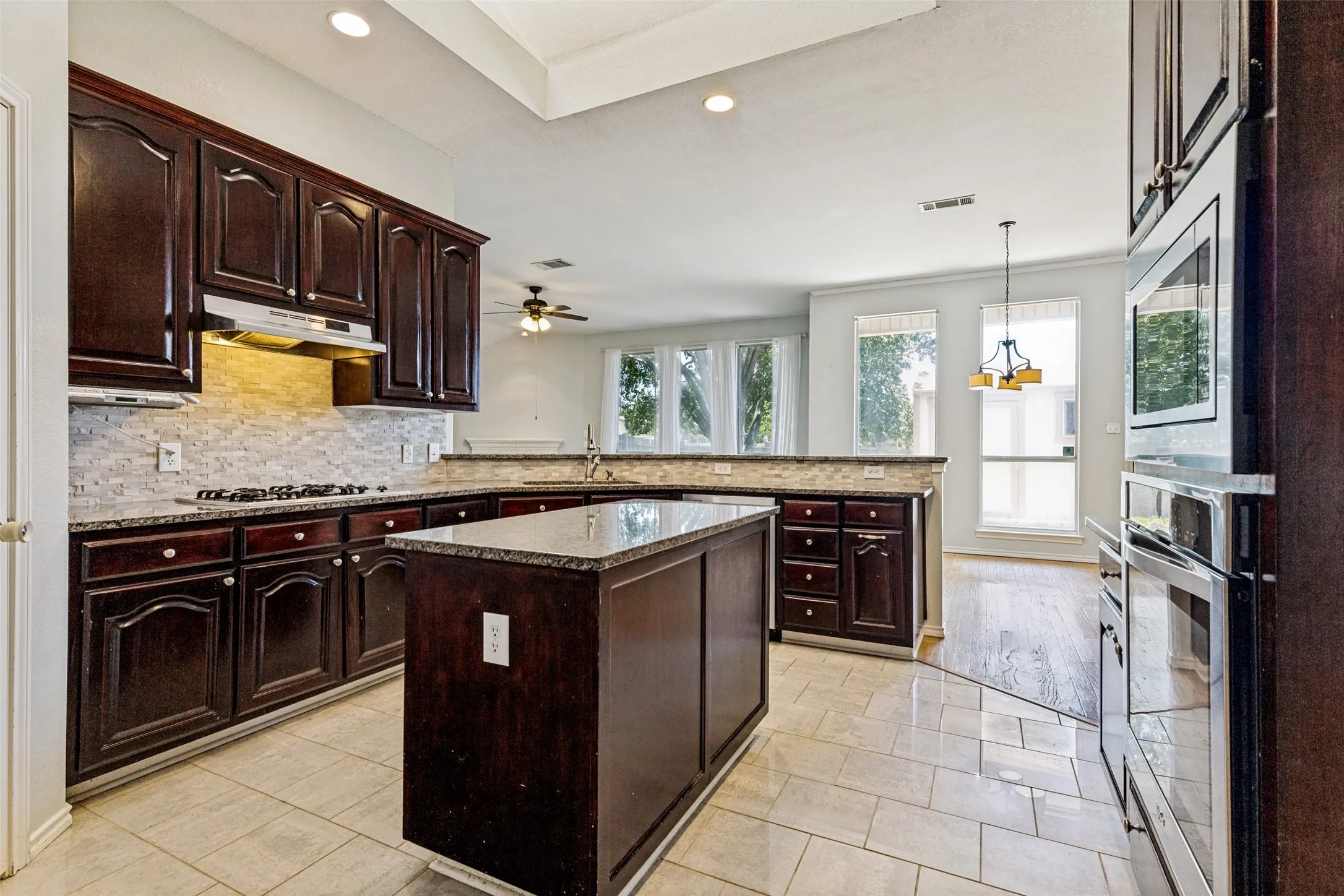 Open-concept kitchen featuring dark wood cabinetry, light granite countertops, a tiled backsplash, and stainless steel built-in appliances