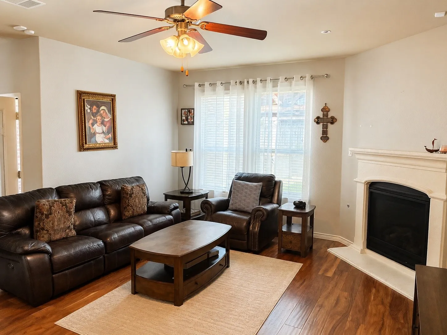 Spacious living area featuring wood-finish flooring, a ceiling fan with integrated lighting, and a white-mantled fireplace