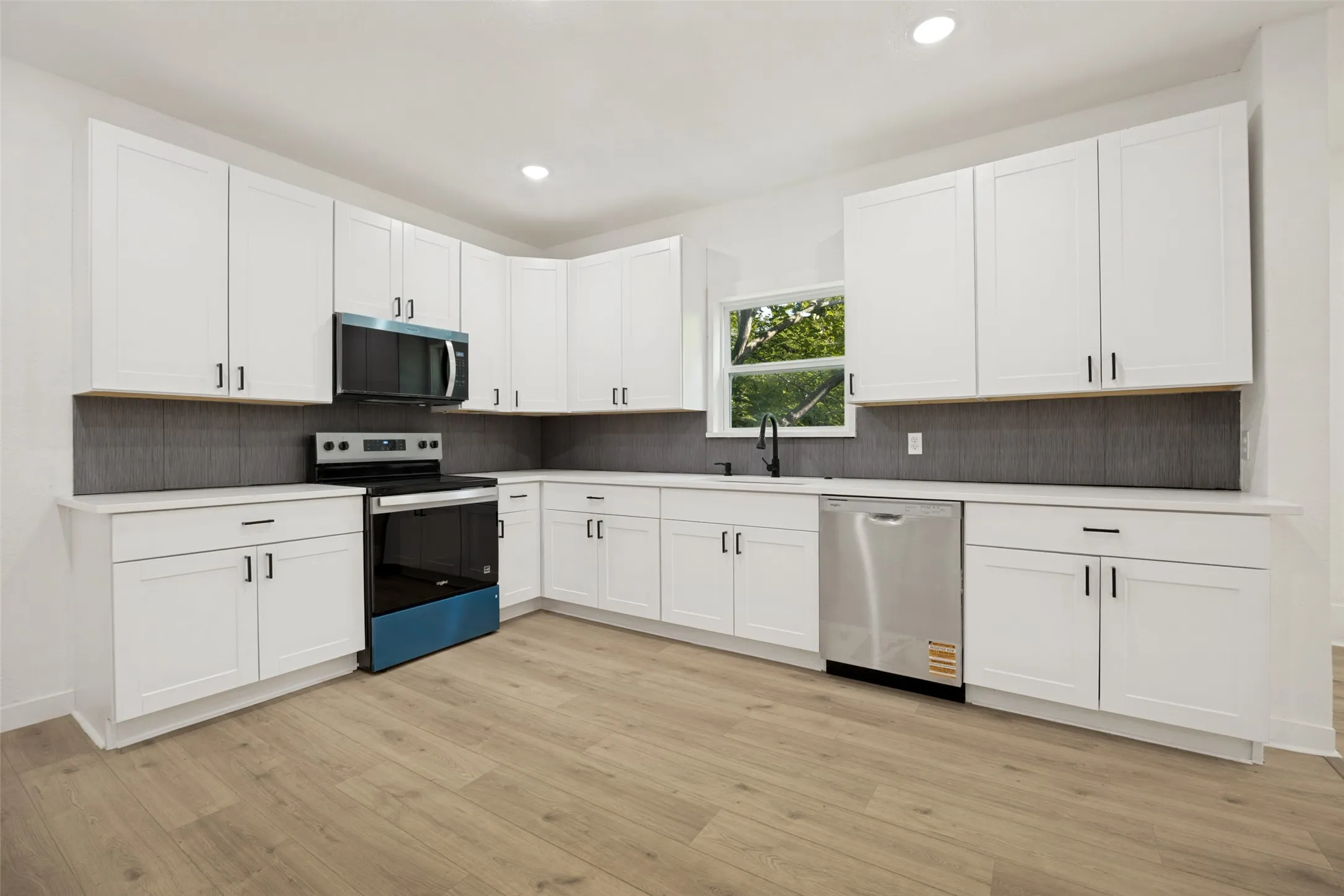 Modern kitchen featuring extensive white cabinetry, light wood-finish flooring, recessed lighting, stainless steel appliances, and a dark tile backsplash