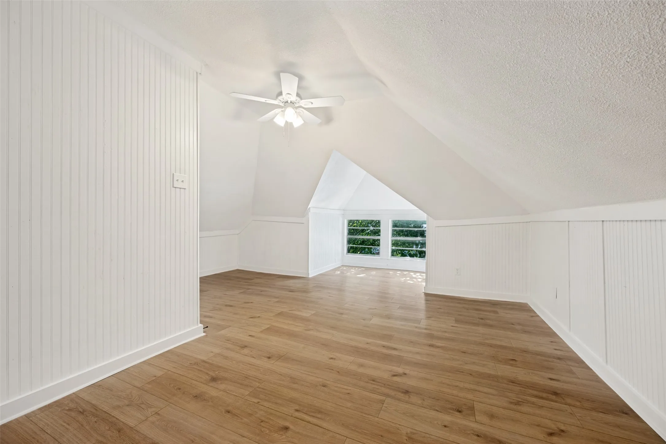 Spacious room featuring light wood-finish flooring, white beadboard paneling, sloped ceilings, and a ceiling fan
