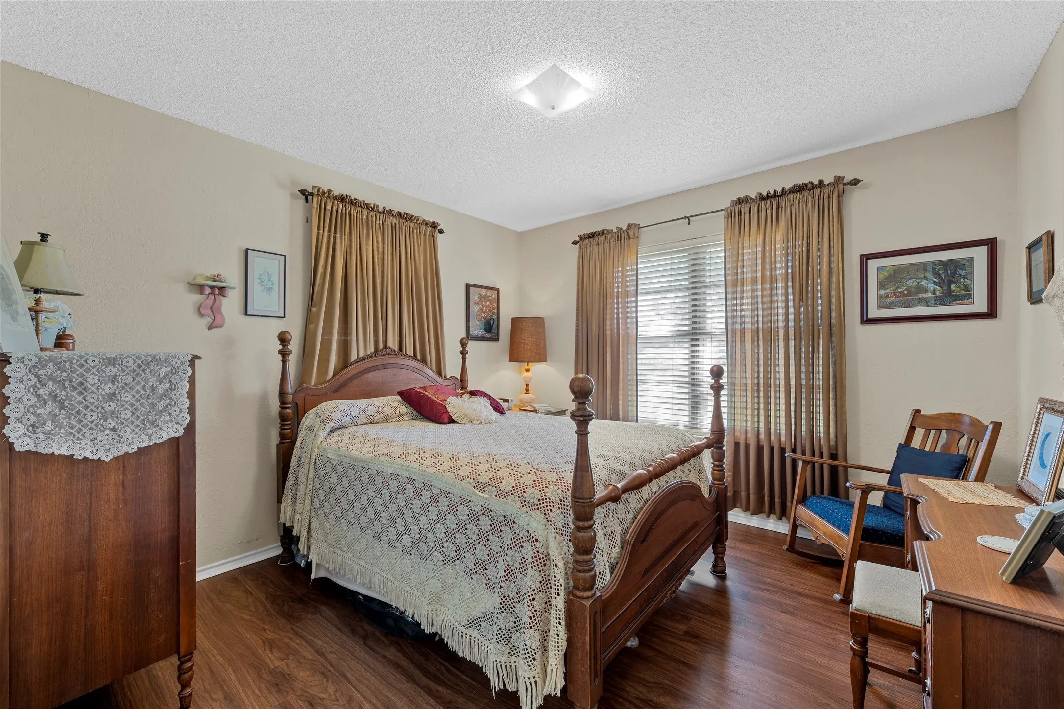 Spacious bedroom featuring wood-finish flooring, light neutral wall tones, window with horizontal blinds and drapery, and a ceiling-mounted light fixture