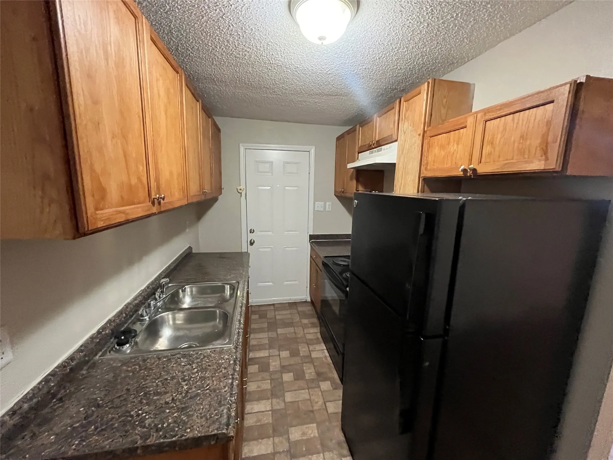 Galley kitchen featuring wood-finish cabinetry, a dual-basin stainless steel sink, dark granite-style countertops, and a black appliance package