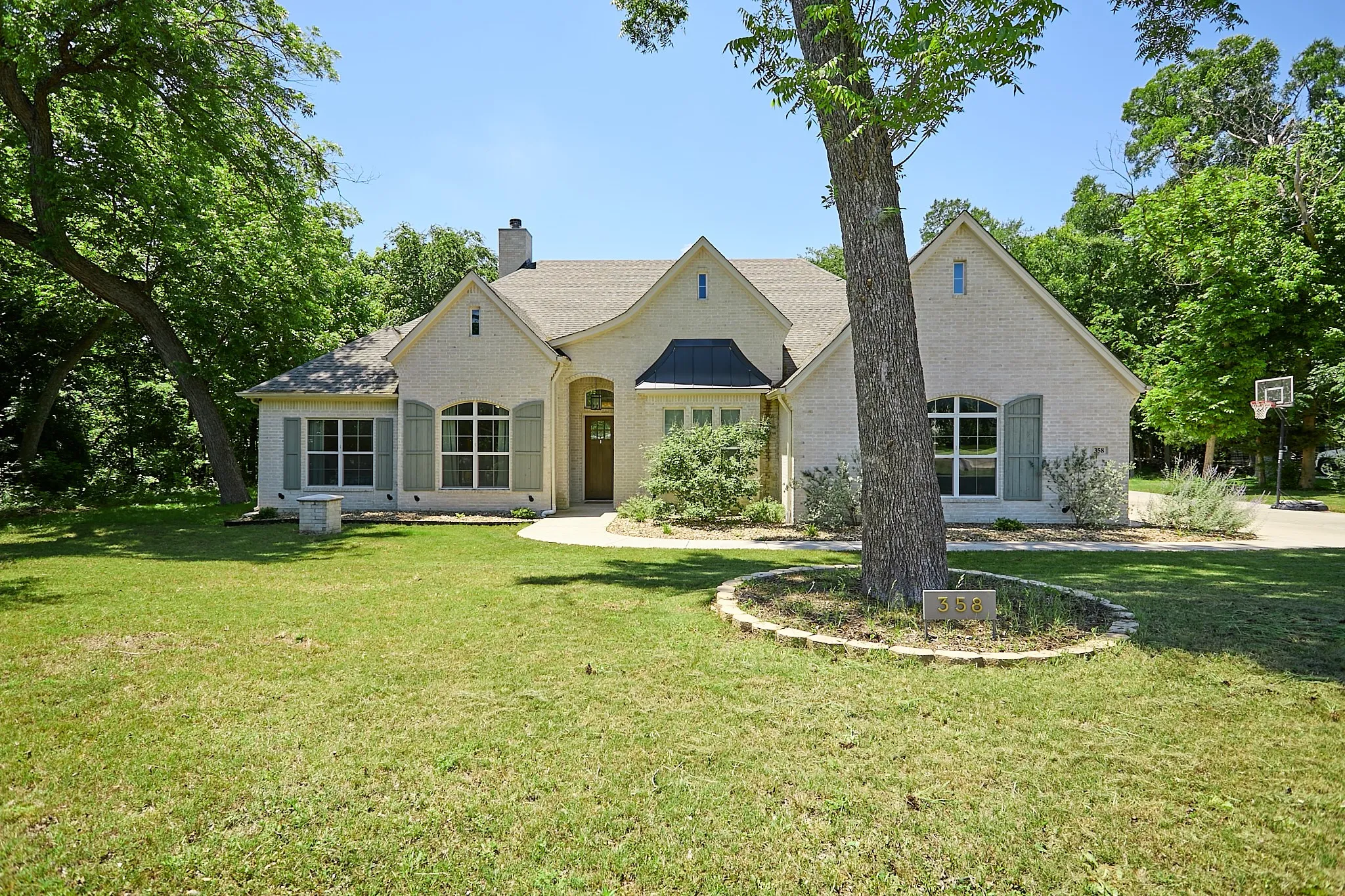 Light-colored brick exterior featuring arched windows, decorative shutters, a prominent front entry with an arched door, a gabled roofline, and mature trees enhancing the landscaping