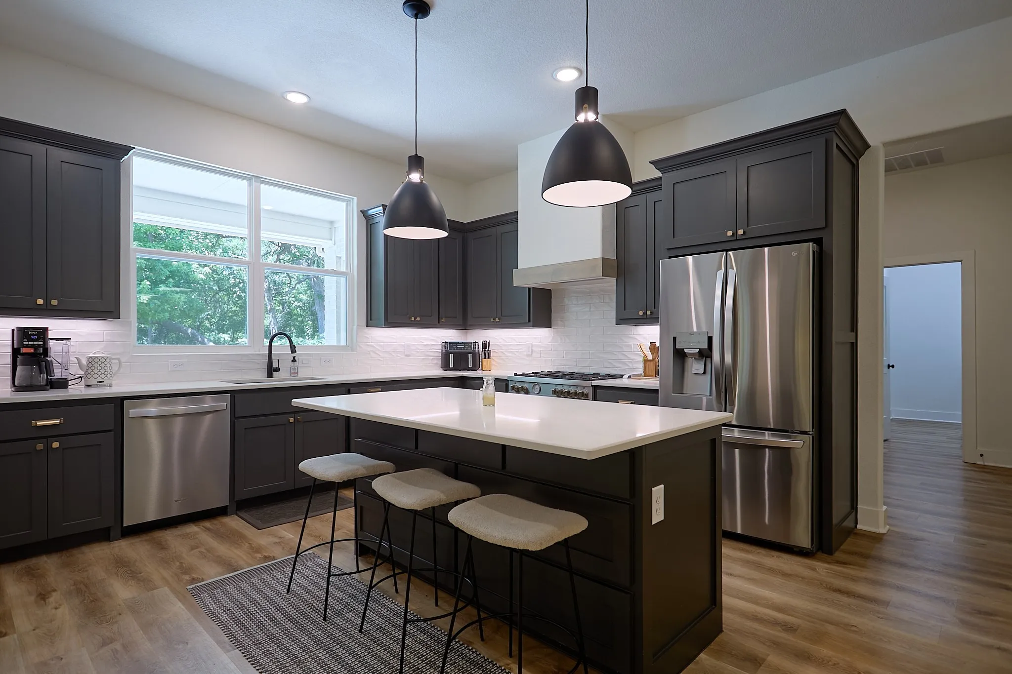 Modern kitchen featuring dark cabinetry with gold-tone hardware, stainless steel appliances, a large center island with a white countertop, and wood-finish flooring