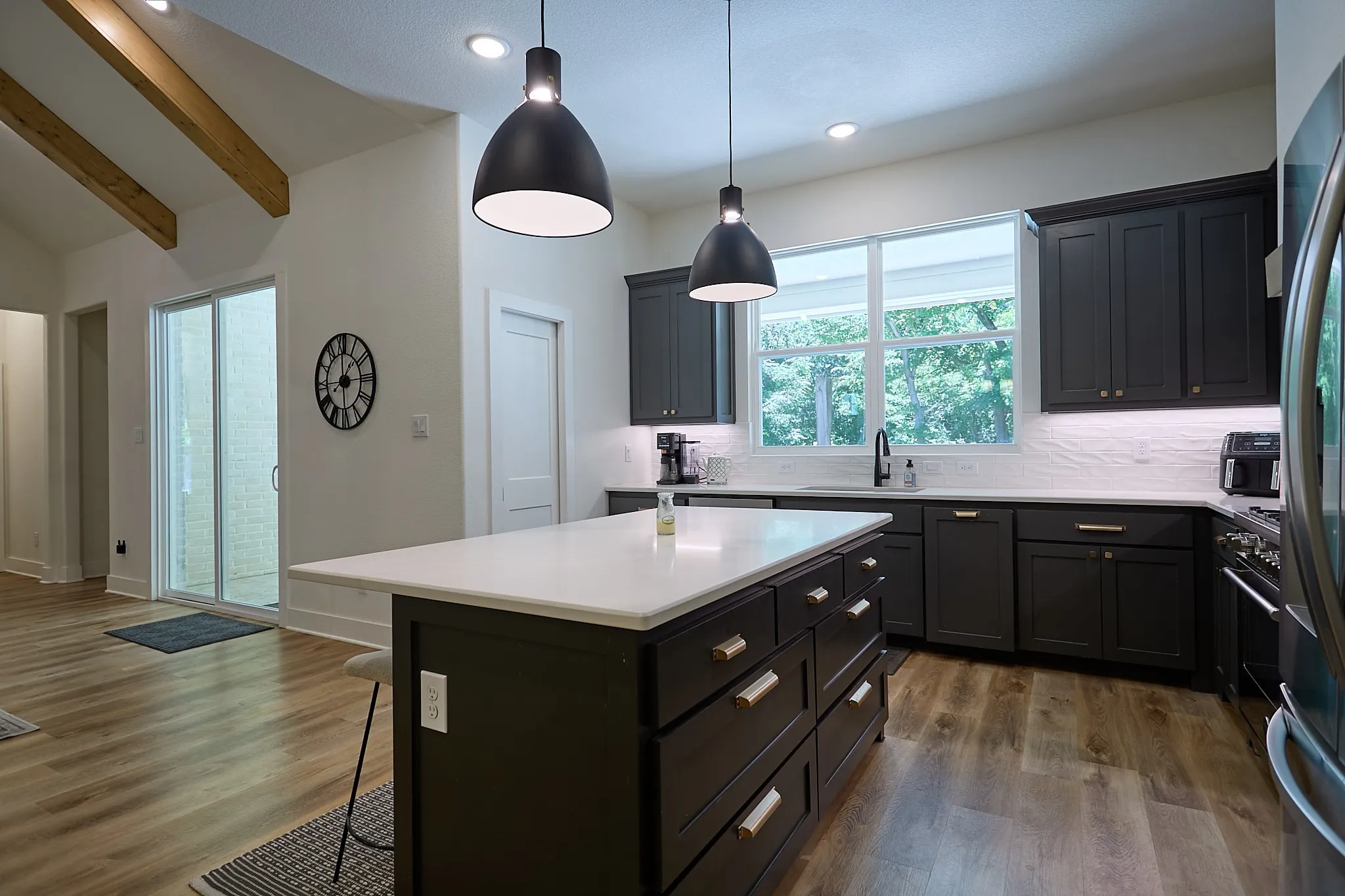 Modern kitchen featuring a large central island with a light-toned countertop, dark cabinetry with gold-toned hardware, wood-finish flooring, and a subway tile backsplash