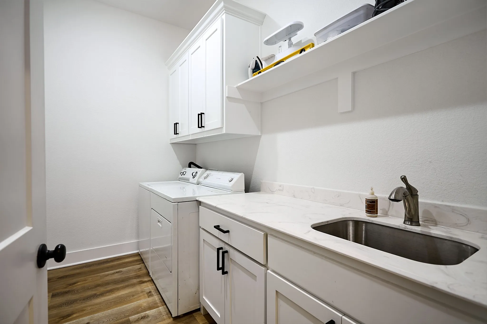 Dedicated laundry room featuring wood-finish flooring, white cabinetry with black hardware, a stainless steel sink, and light-toned countertops
