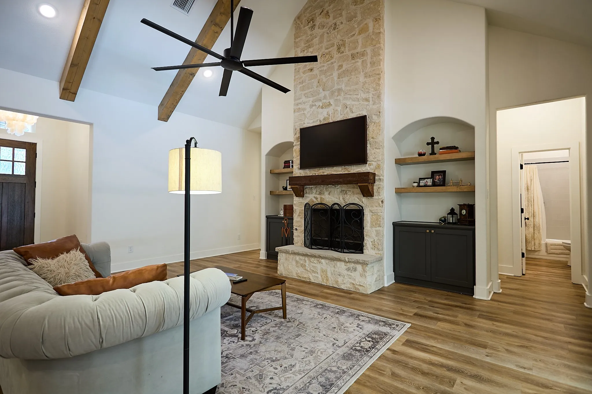 Living area featuring a stone fireplace with a wood mantle, vaulted ceiling with exposed wood beams, wood-finish flooring, and built-in shelving with lower cabinetry