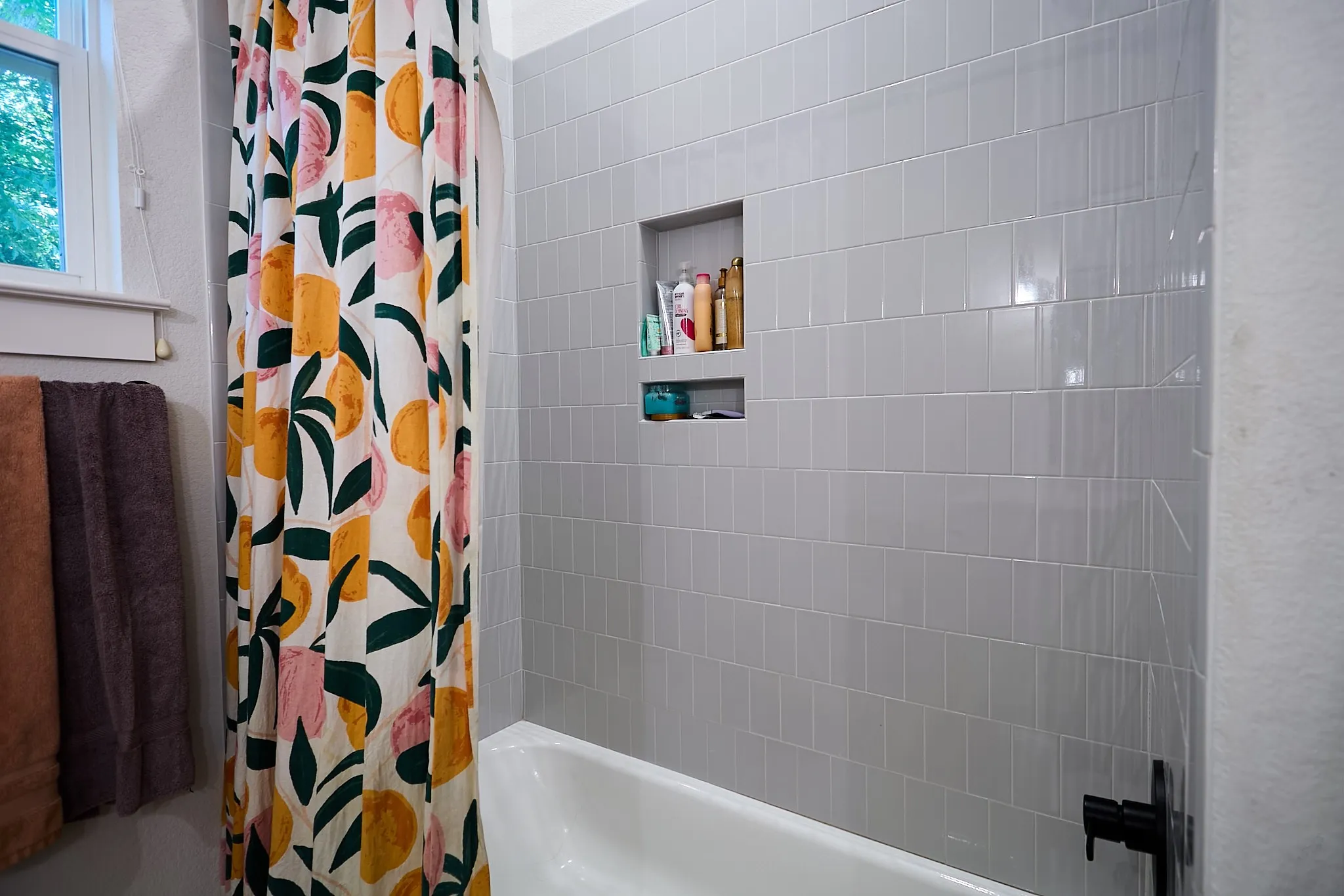 White bathtub with light gray subway tile surround featuring a built-in shower niche and matte black fixtures