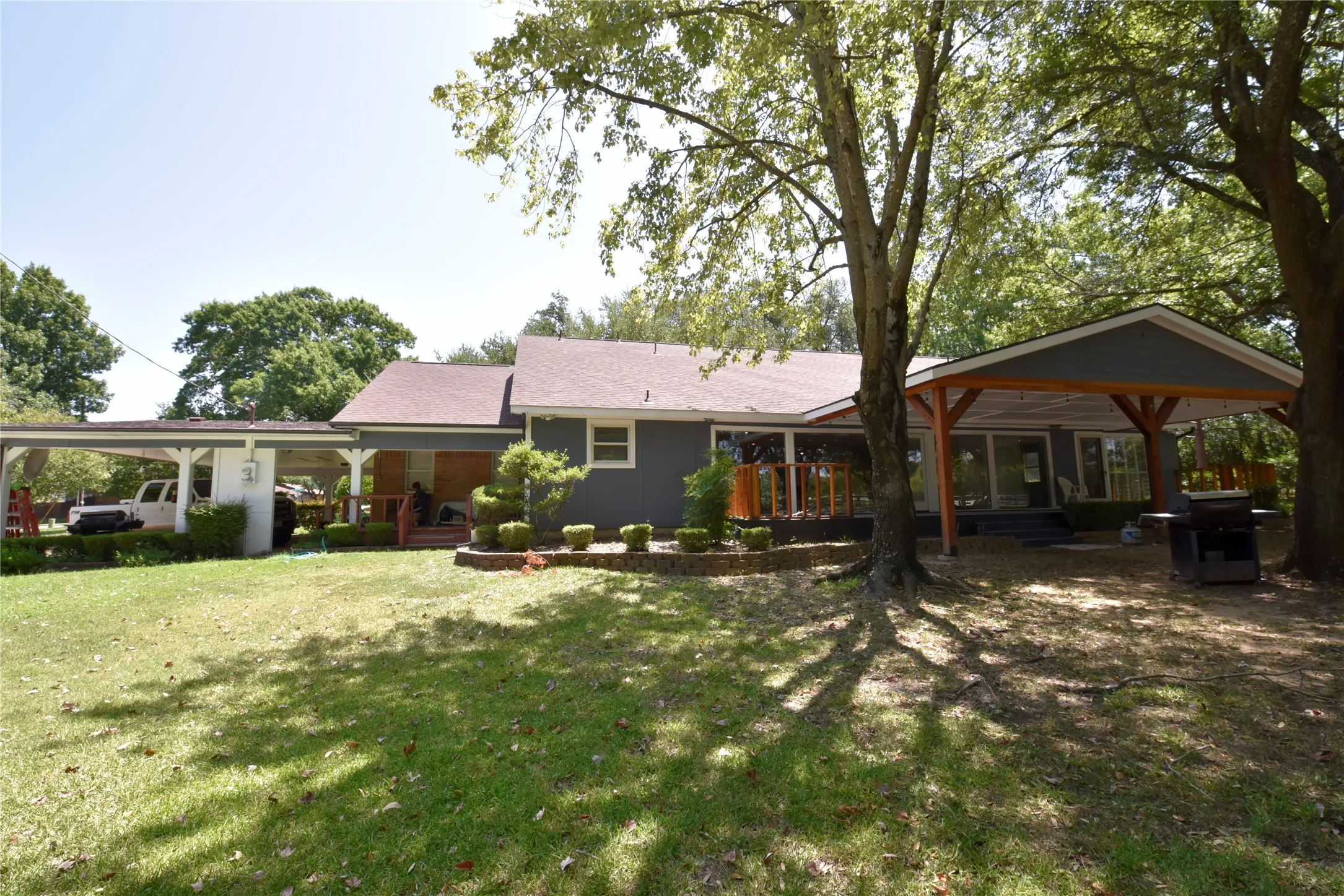 Expansive rear yard featuring a covered patio with exposed wood beams, a covered carport, and mature trees providing natural shade