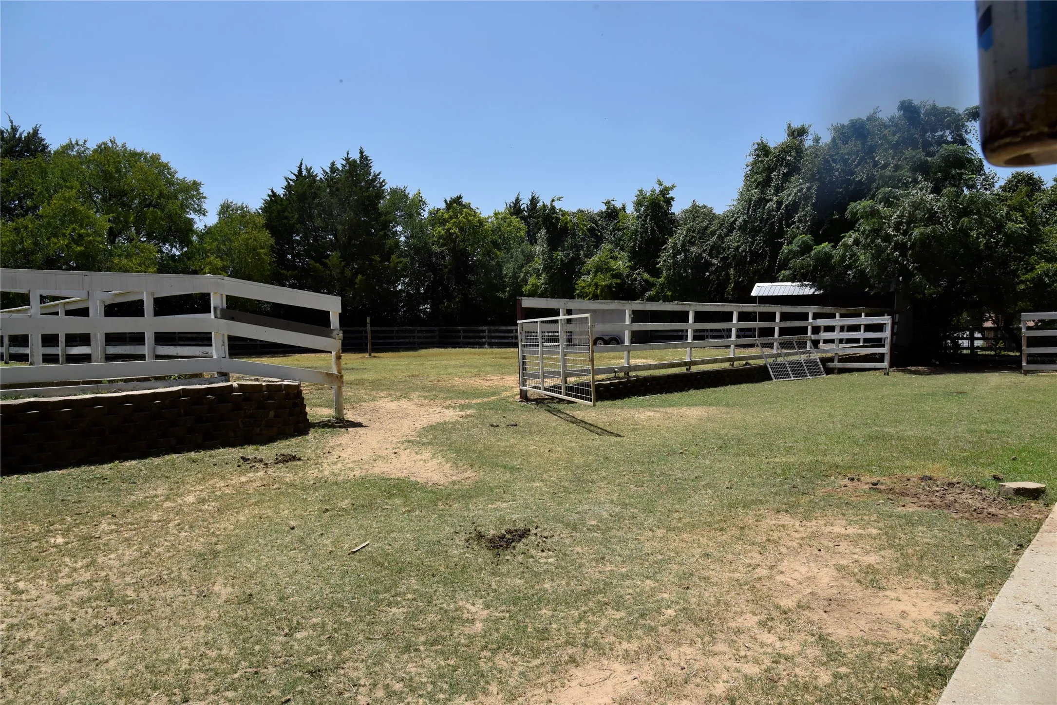 Expansive outdoor area featuring white ranch-style fencing, a metal gate, and mature perimeter trees