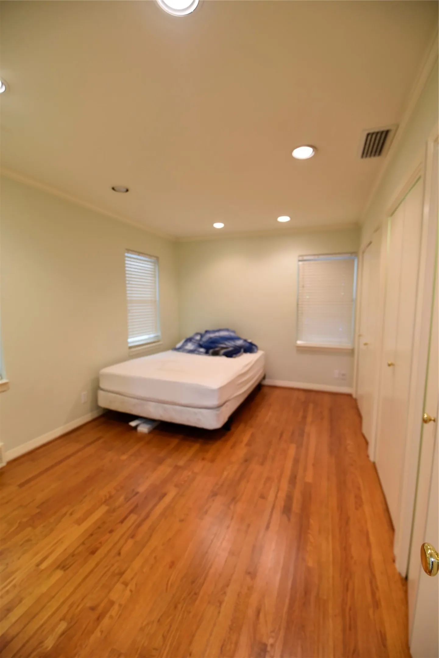 Spacious room featuring wood-finish flooring, recessed ceiling lighting, two windows with blinds, and two paneled doors with brass-toned hardware