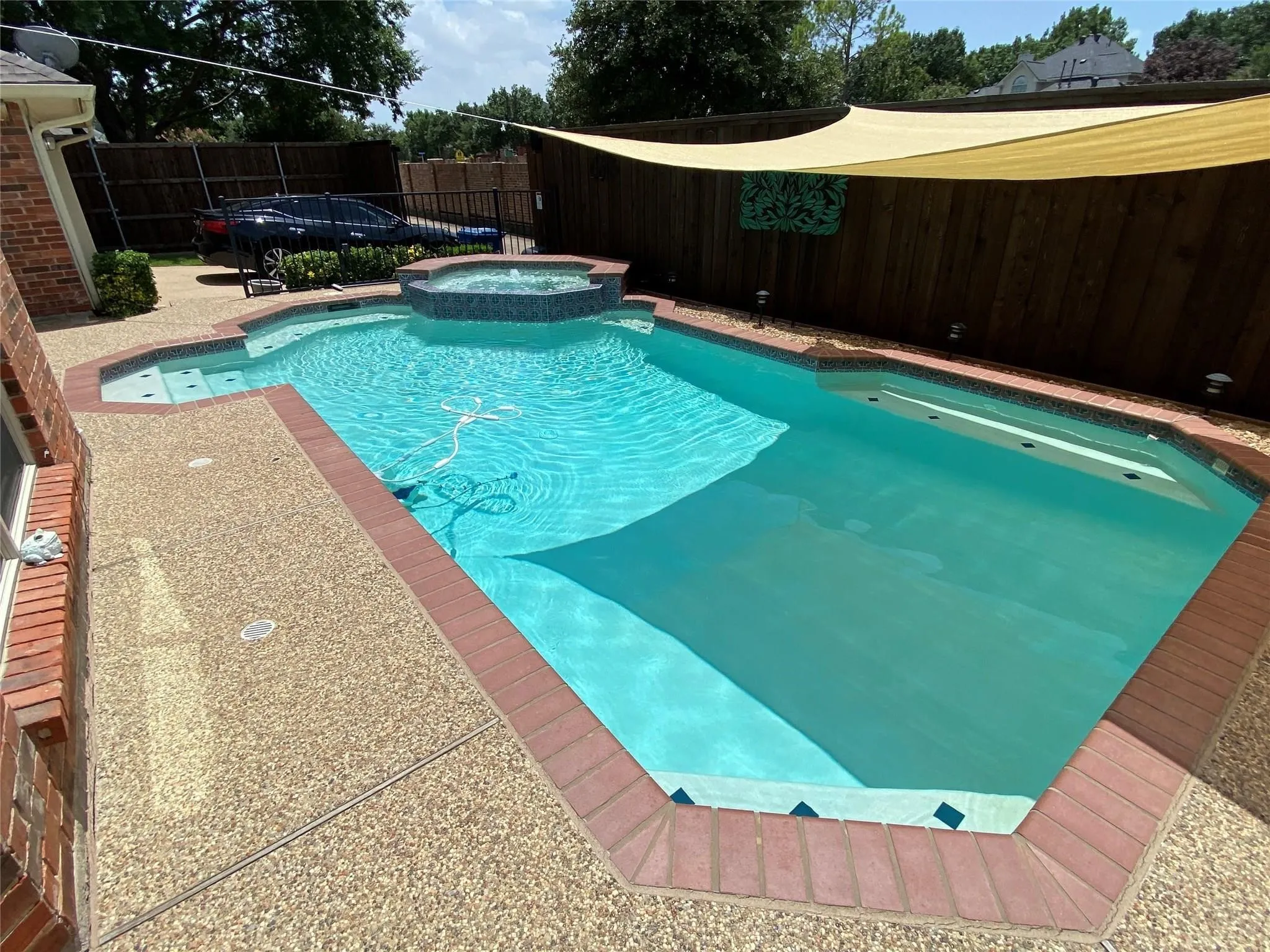 Freeform swimming pool featuring an integrated spa, exposed aggregate decking, brick coping, and a privacy fence