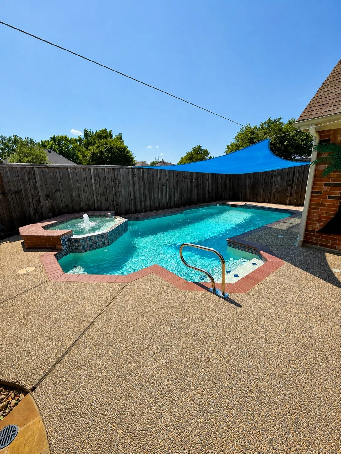 Custom-shaped swimming pool with an integrated spa featuring a water fountain, surrounded by exposed aggregate concrete decking and a privacy fence