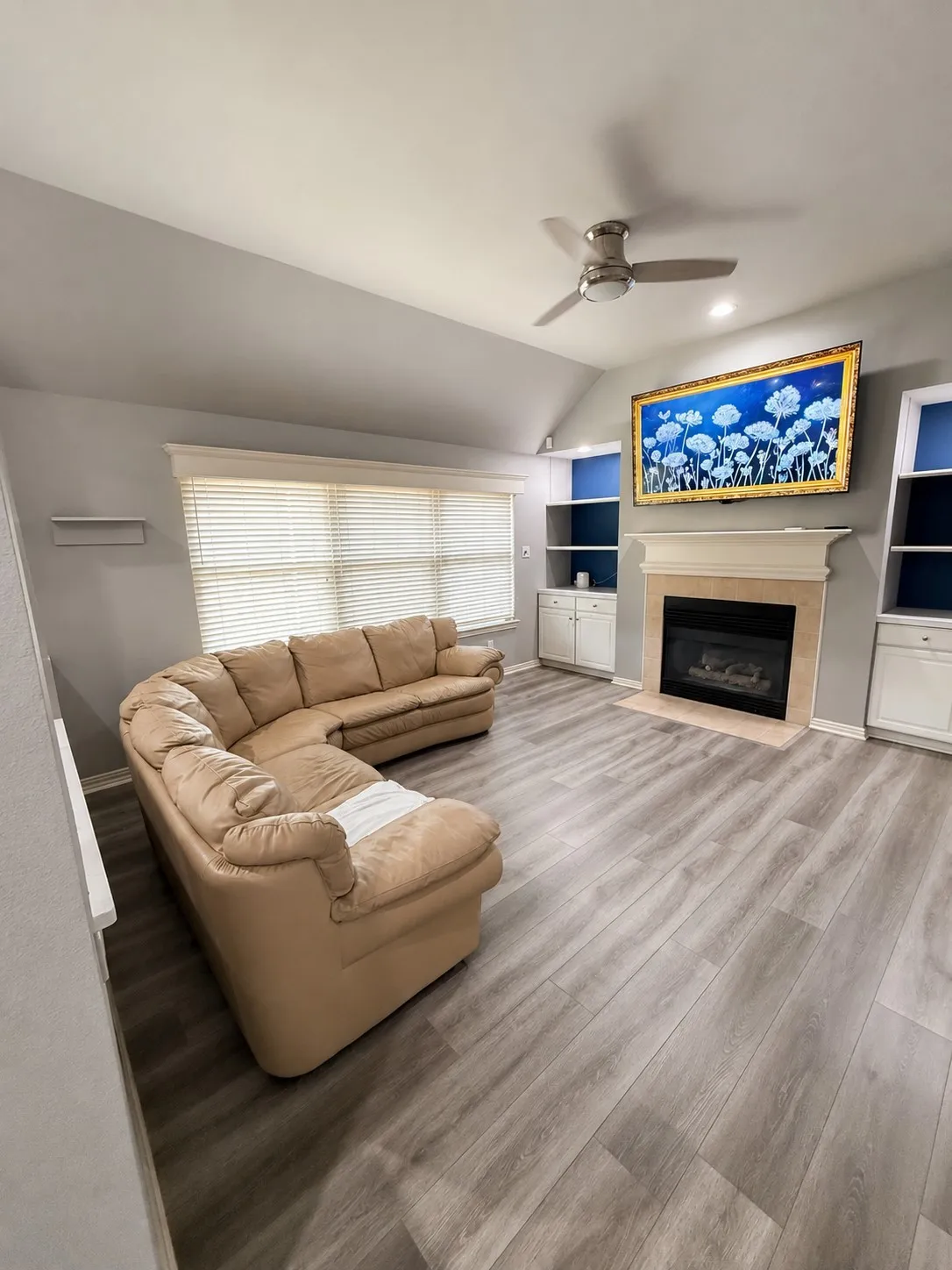 Vaulted ceiling with a ceiling fan, wood-finish flooring, built-in shelving, a fireplace with a mantel, and a large window with blinds